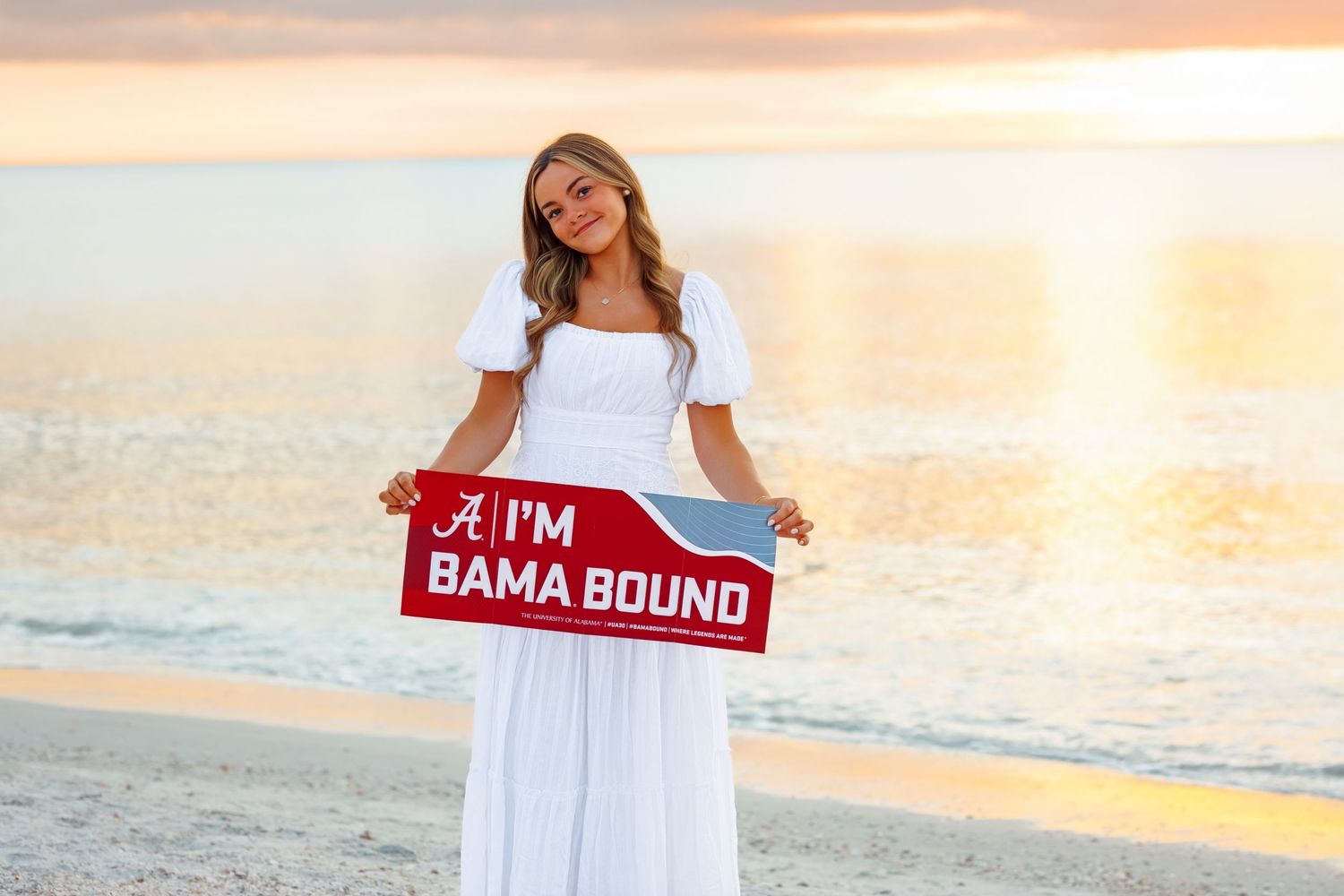 Young woman in white dress holding an I'm Bama Bound sign on a beach at sunset.