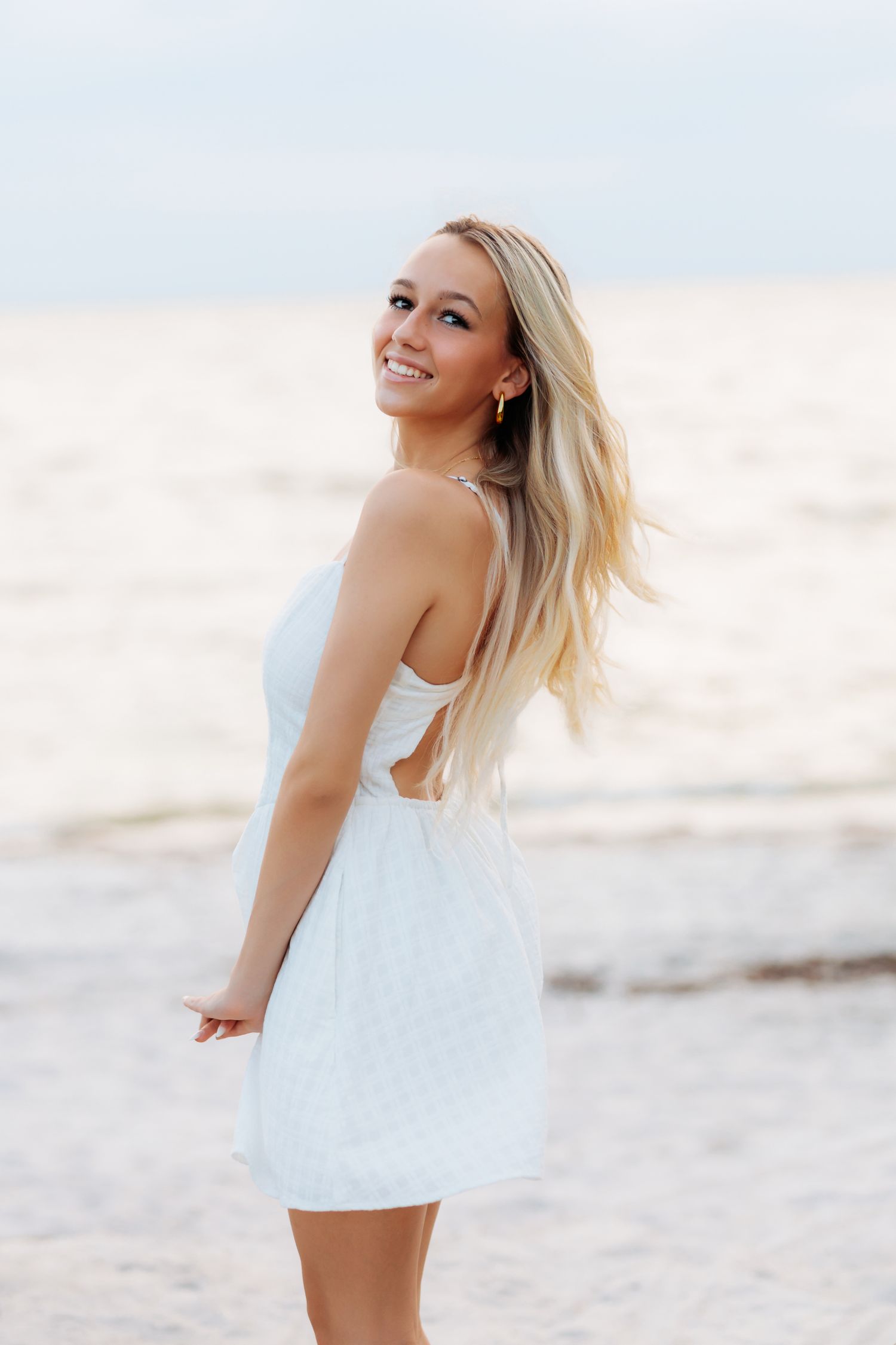 high school senior girl in white dress smiling at beach, hair flowing in breeze, ocean backdrop.