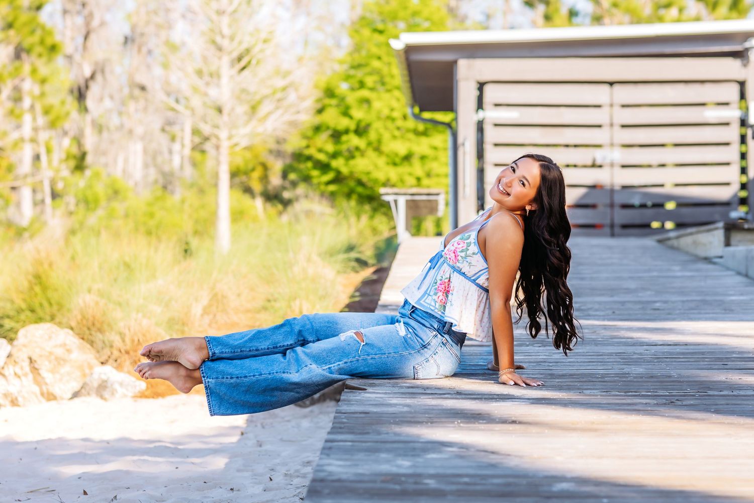 Young woman with long dark hair poses on wooden boardwalk in jeans and floral top, surrounded by lush greenery.