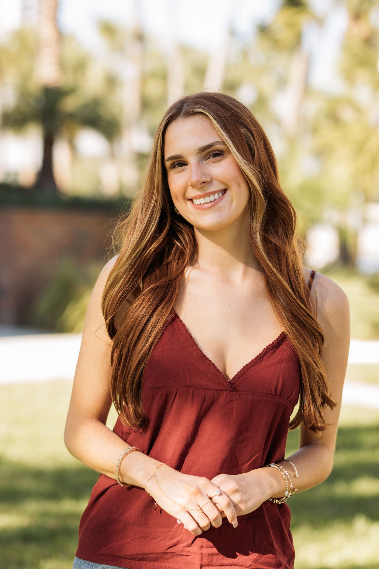 Smiling young woman with long auburn hair wearing a red sleeveless top, posing outdoors with palm trees in background.