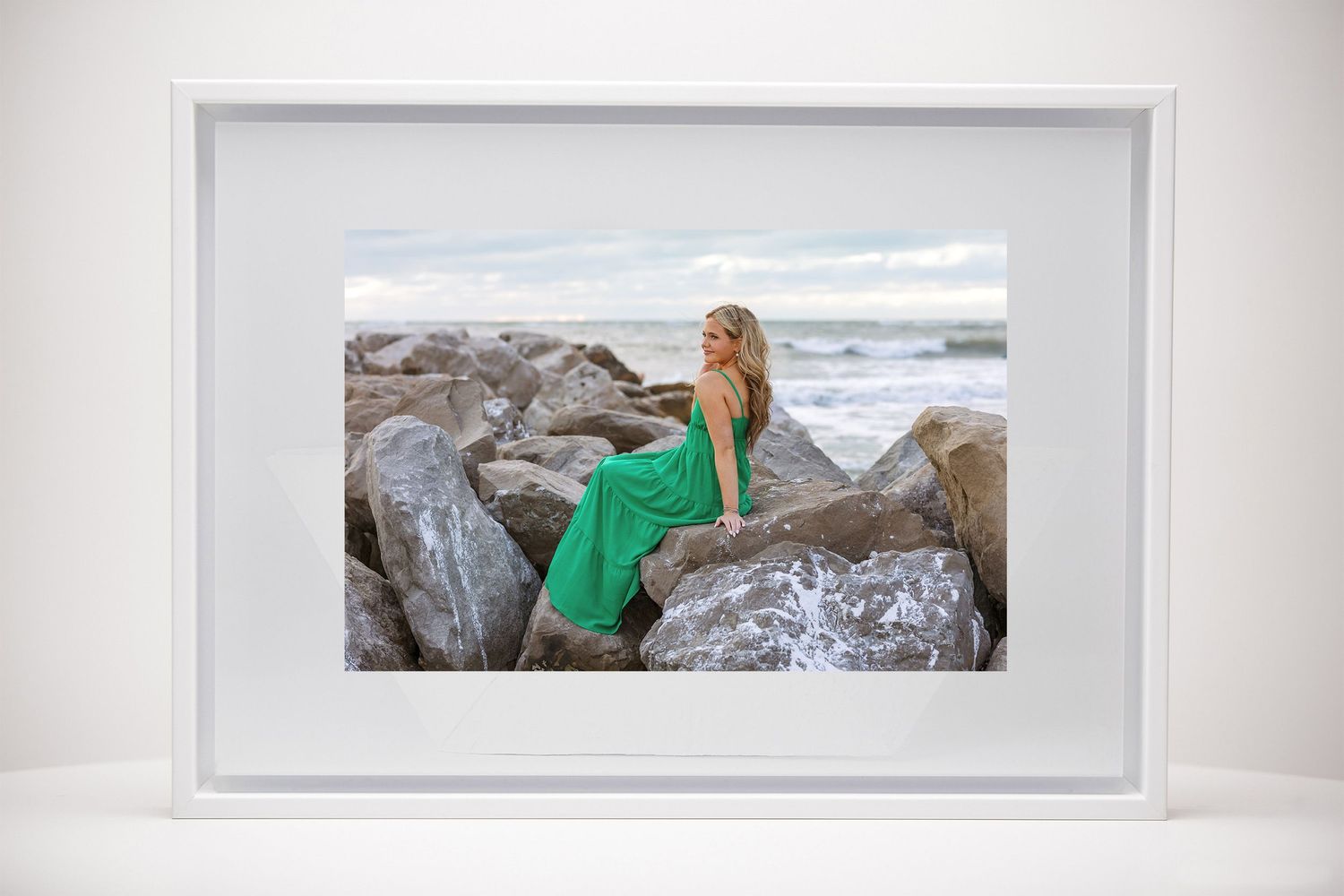 Framed photo of blonde woman in green dress on rocky beach displayed on white surface.
