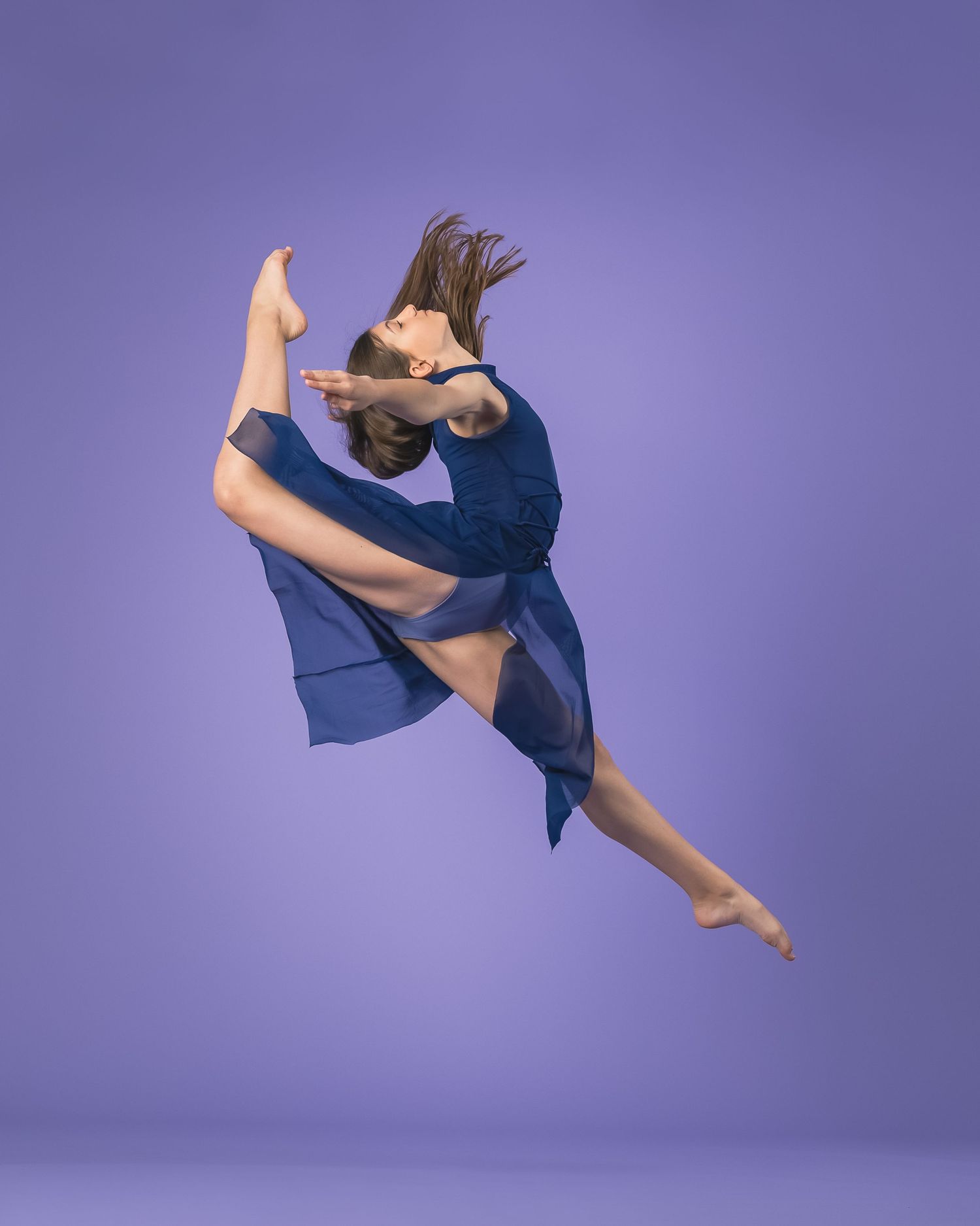 Dancer leaps in blue flowing dress against purple studio background with dynamic mid-air pose.