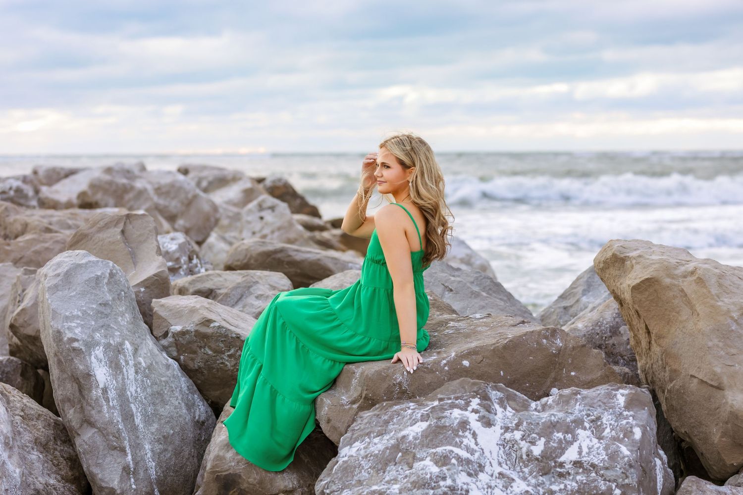 Blonde woman in a flowing green dress sits on coastal rocks with ocean waves in the background.