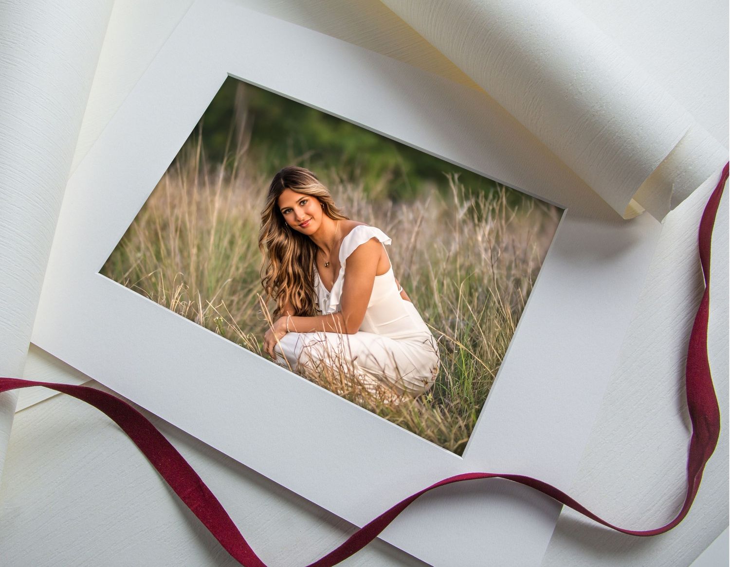A senior portrait print of a young woman sitting in a field, displayed in a white mat with ribbon.