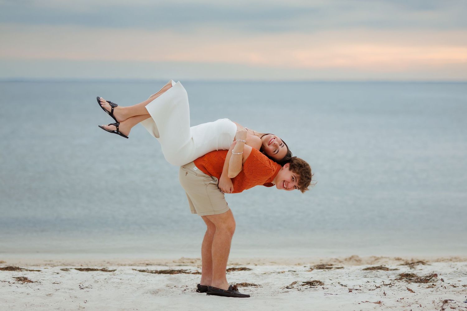 A man lifts a woman in a white dress on a sandy beach with calm ocean waters in the background.