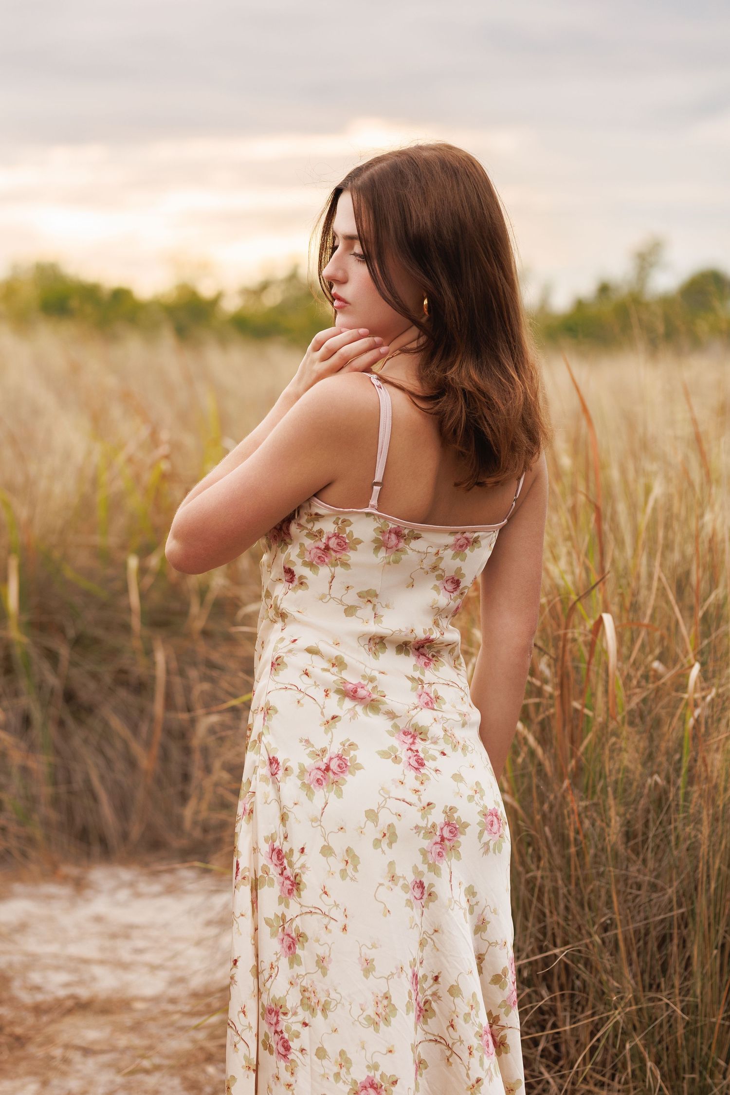 Brunette woman in floral sundress standing in golden field at sunset.