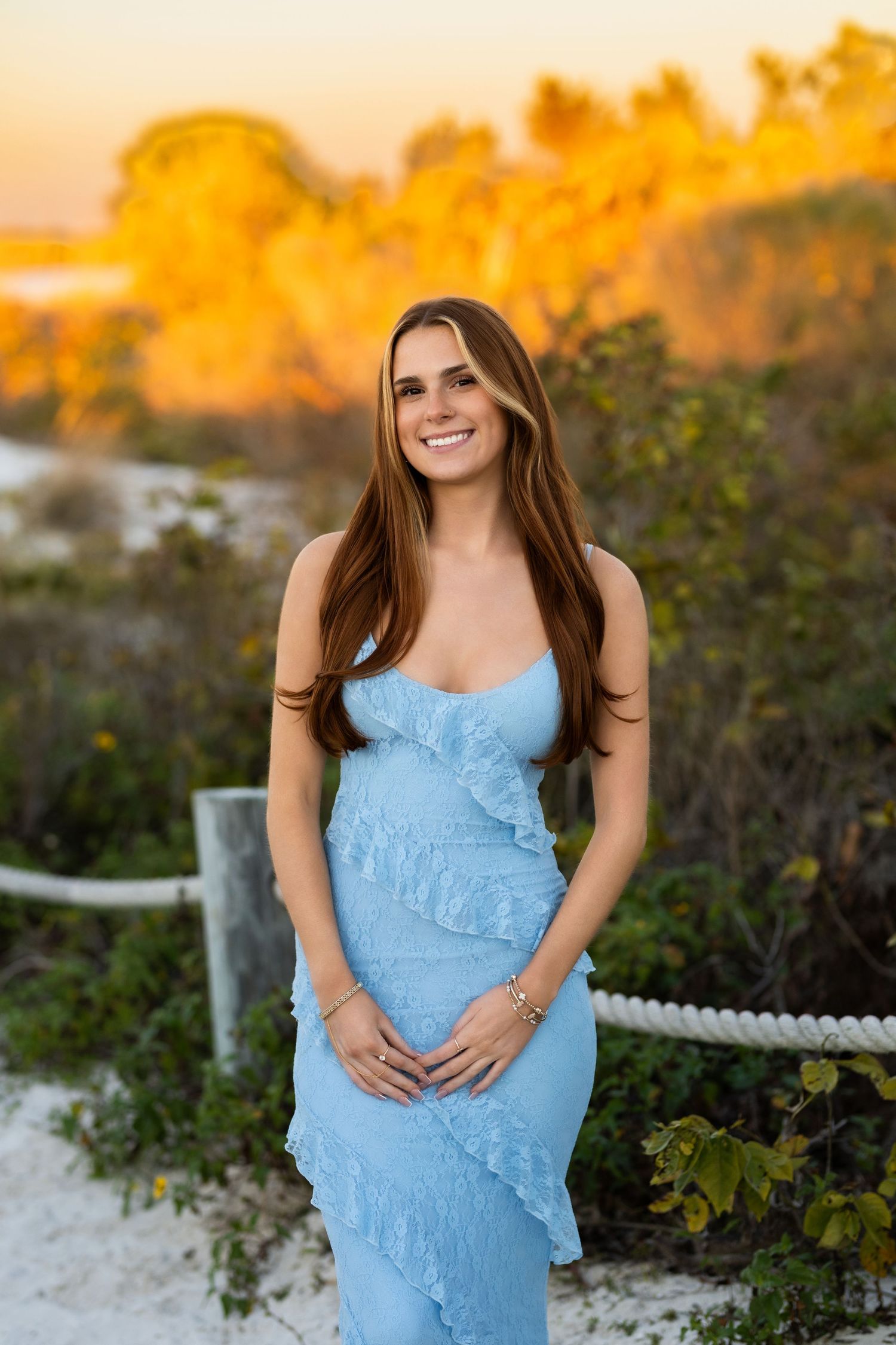 Young woman in blue ruffled dress smiling at sunset on a sandy beach path.
