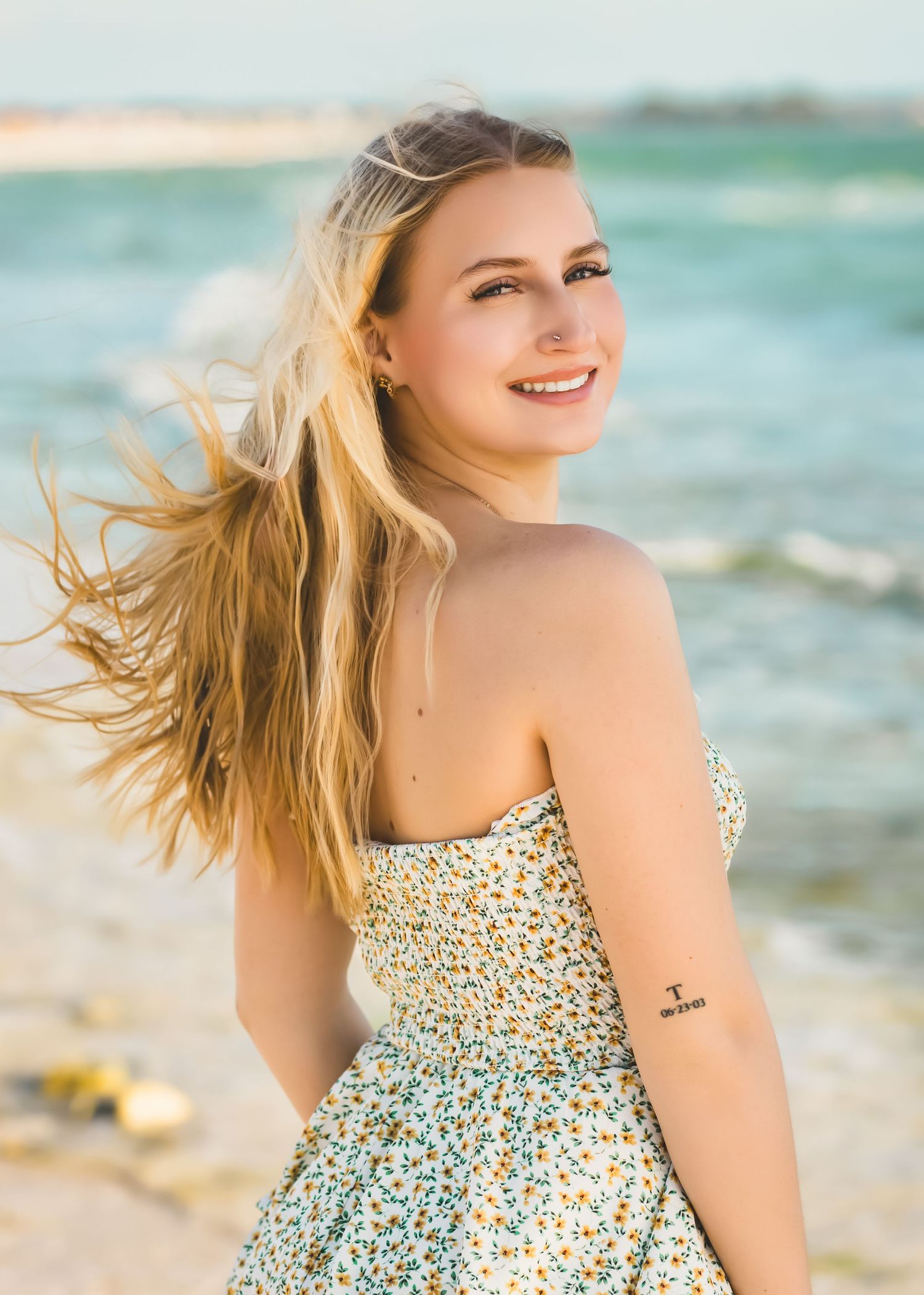 Smiling blonde woman in floral sundress with wind-blown hair at sunny beach shoreline.