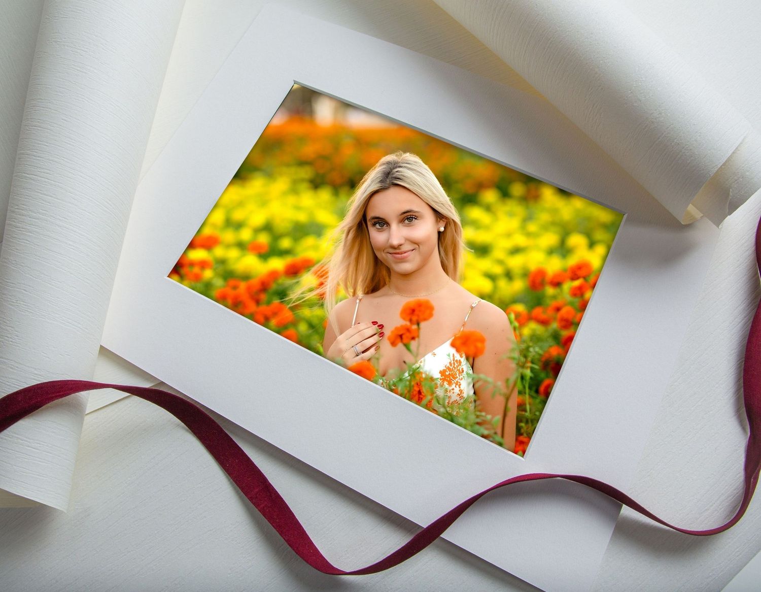 A blonde senior portrait photo of a girl in a flower field displayed in an elegant white folio with ribbon.