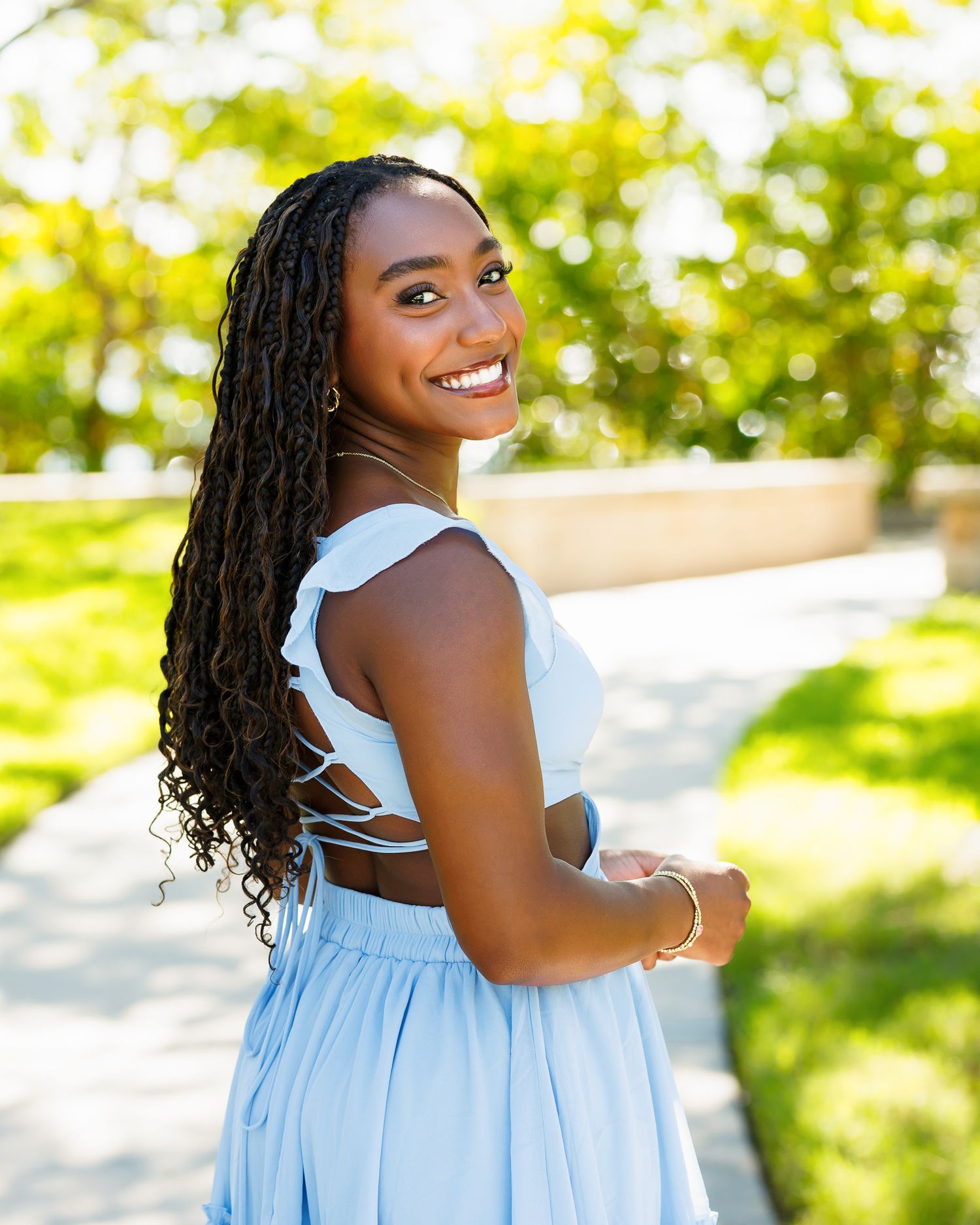 Young Black woman with long braids smiles over shoulder in light blue dress outdoors on sunny day.