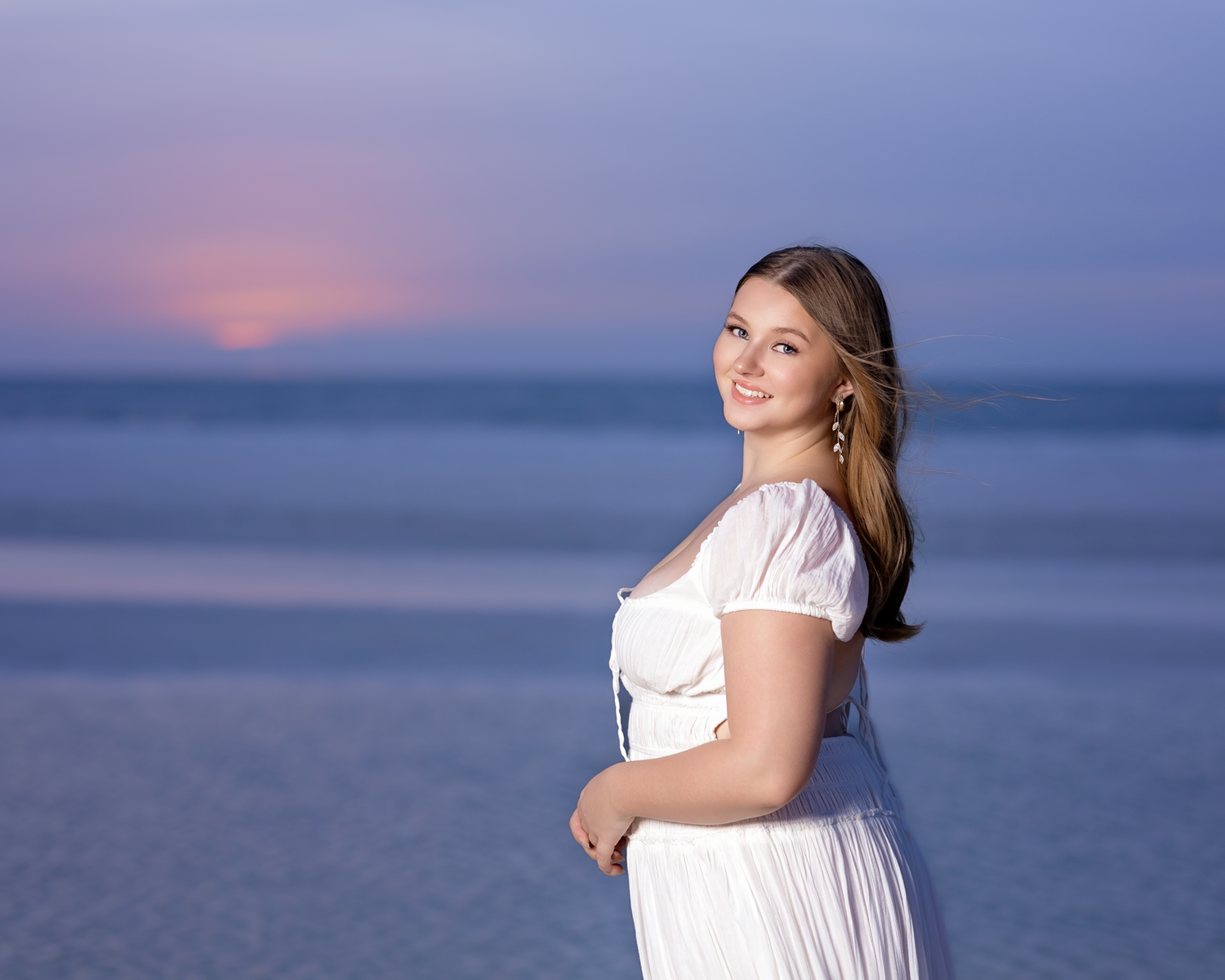 Smiling woman in white dress posing on beach at dusk with colorful sunset sky.
