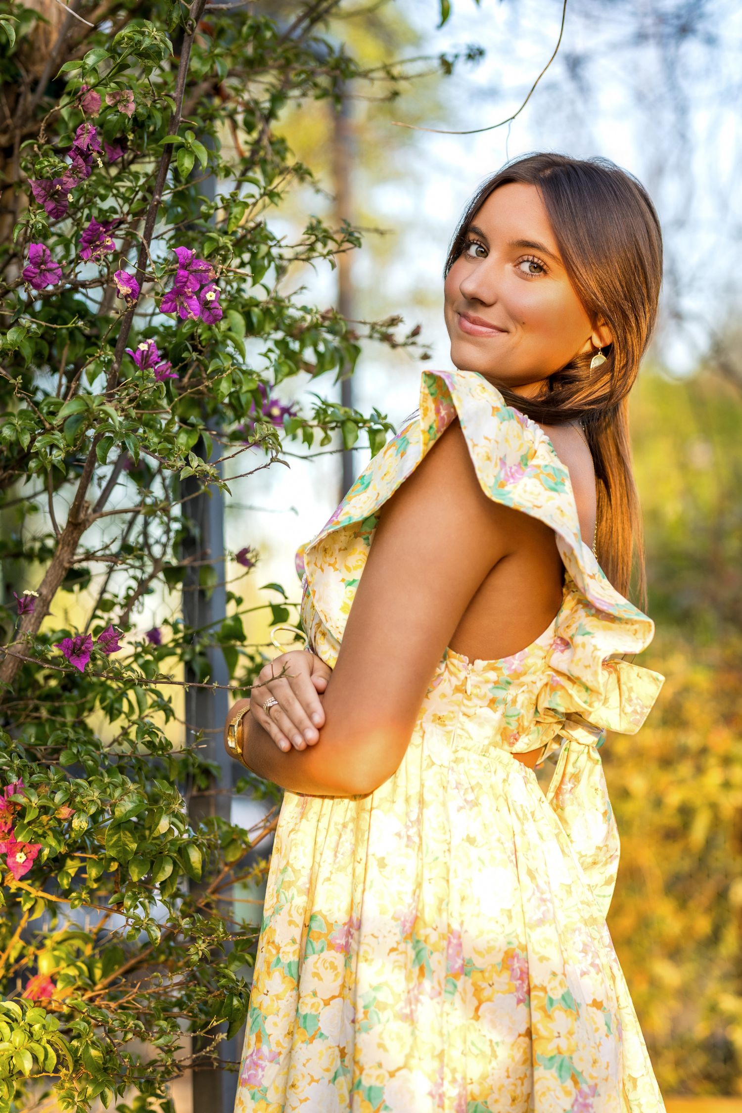 Young woman in a yellow floral backless dress stands near blooming purple bougainvillea in a garden.