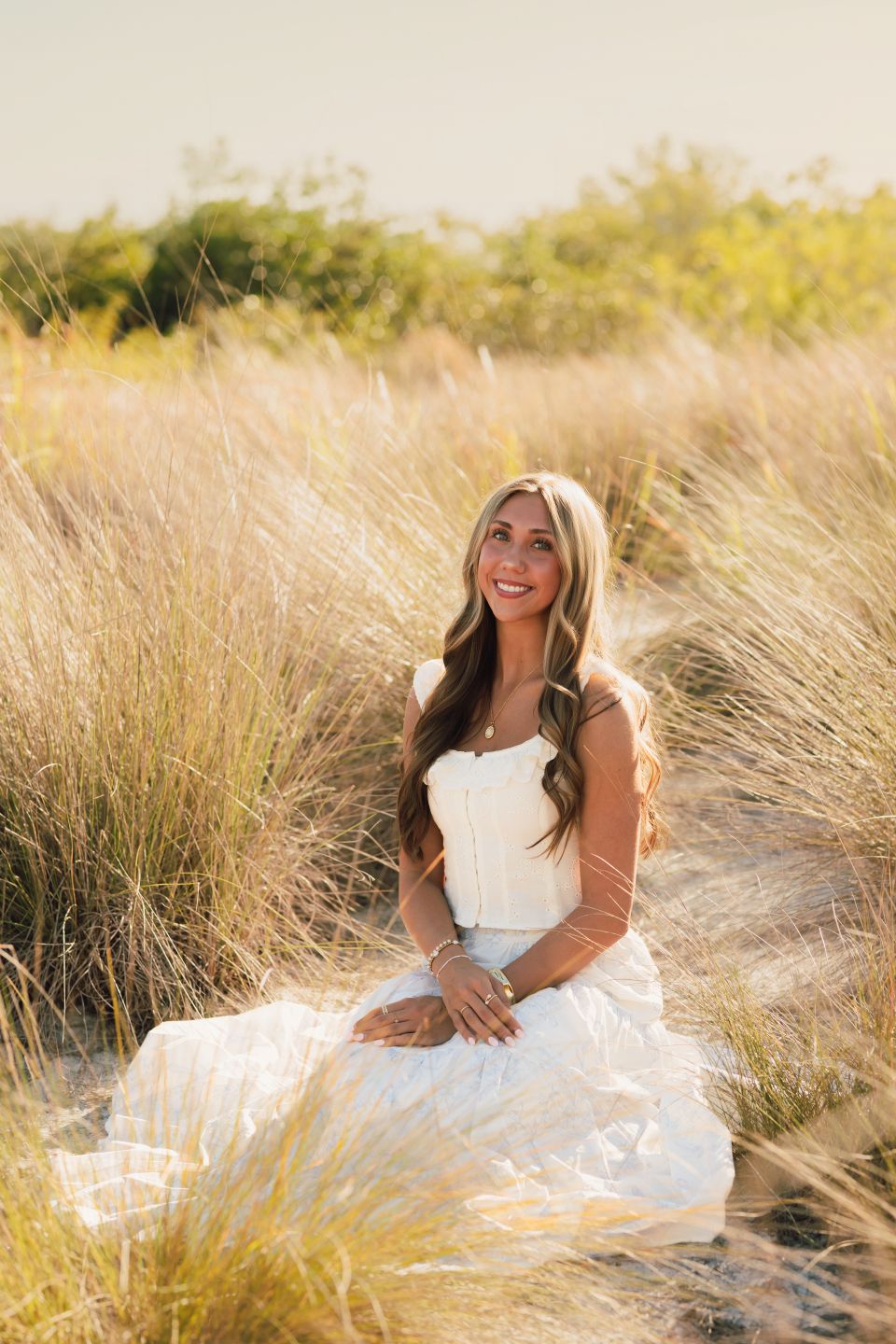Young woman in white dress sitting among golden beach grass dunes, smiling in warm sunset light.