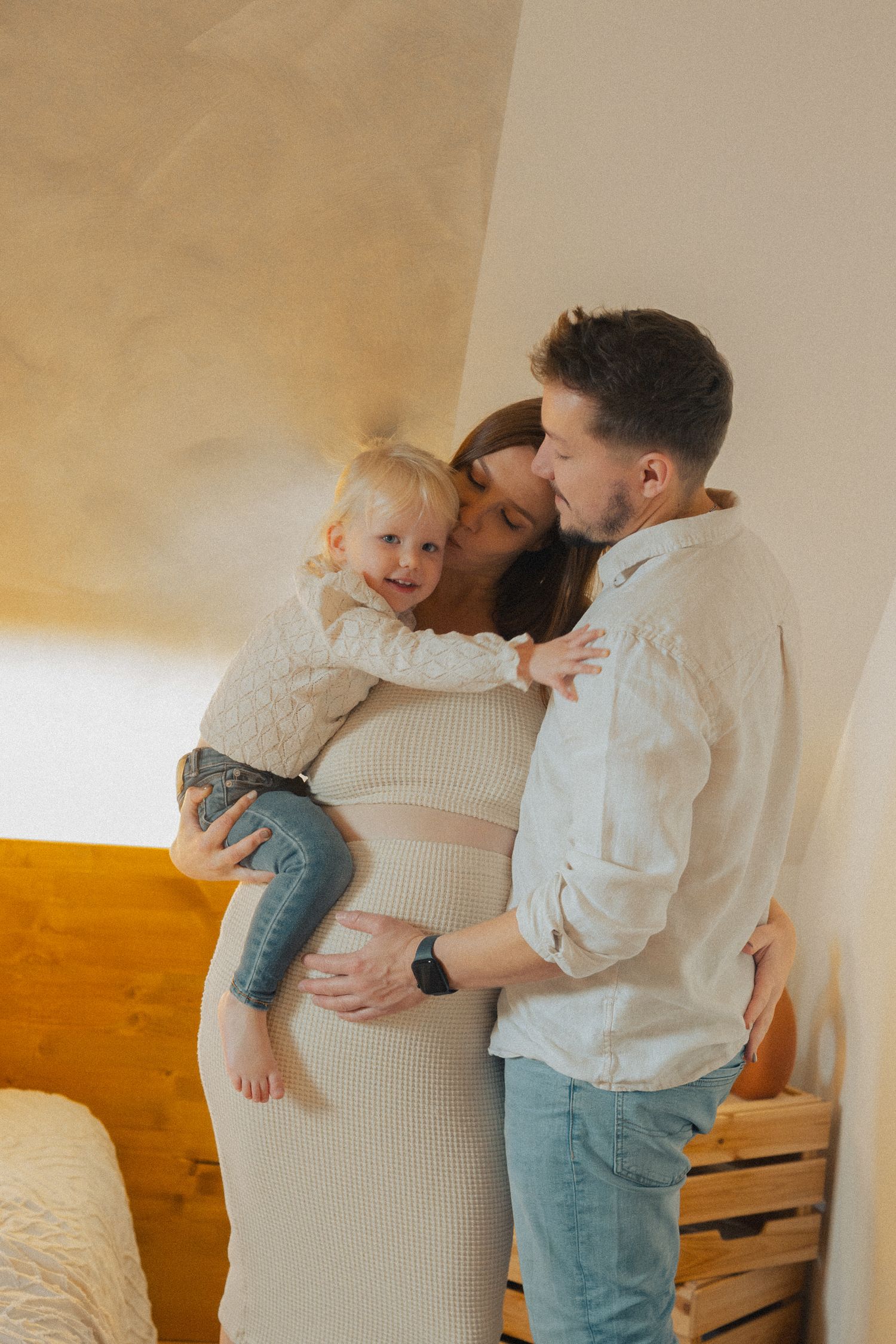 Happy family with pregnant mother kissing father while holding smiling toddler in bedroom.