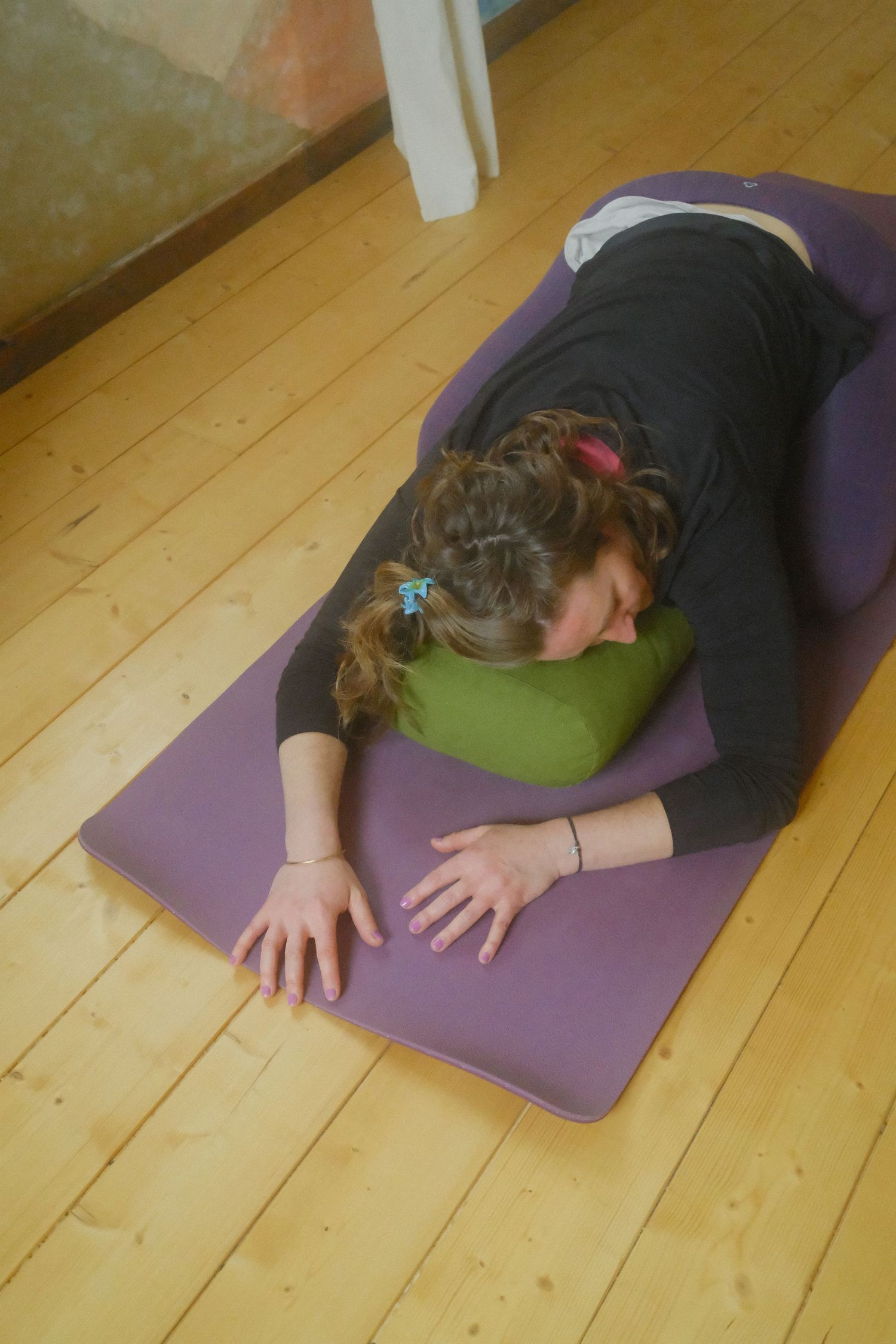 Woman practicing restorative yoga child's pose on purple mat with green bolster on wooden floor.