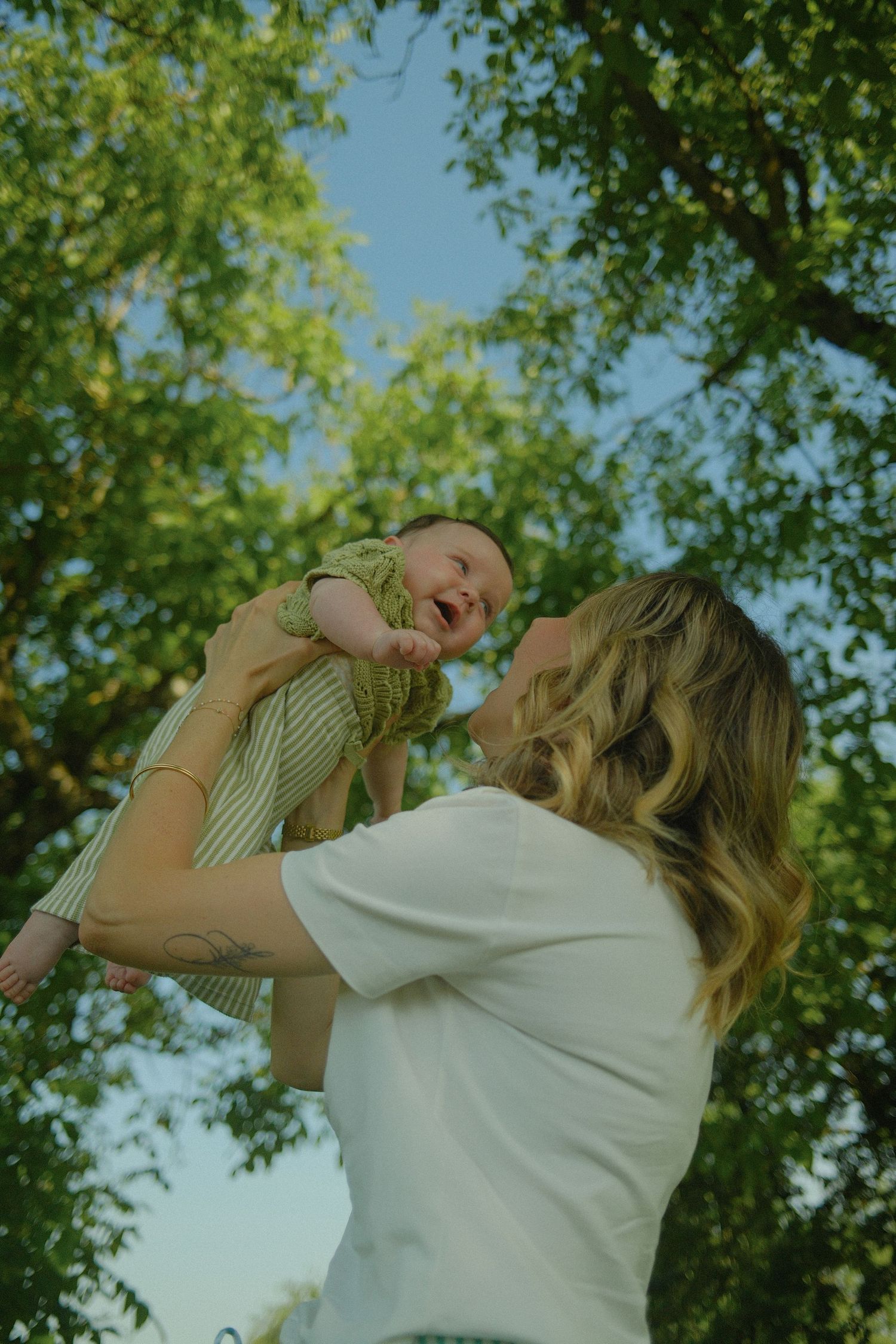 A joyful mother lifts her laughing baby in a green park, sharing a tender moment under lush trees on a sunny day.