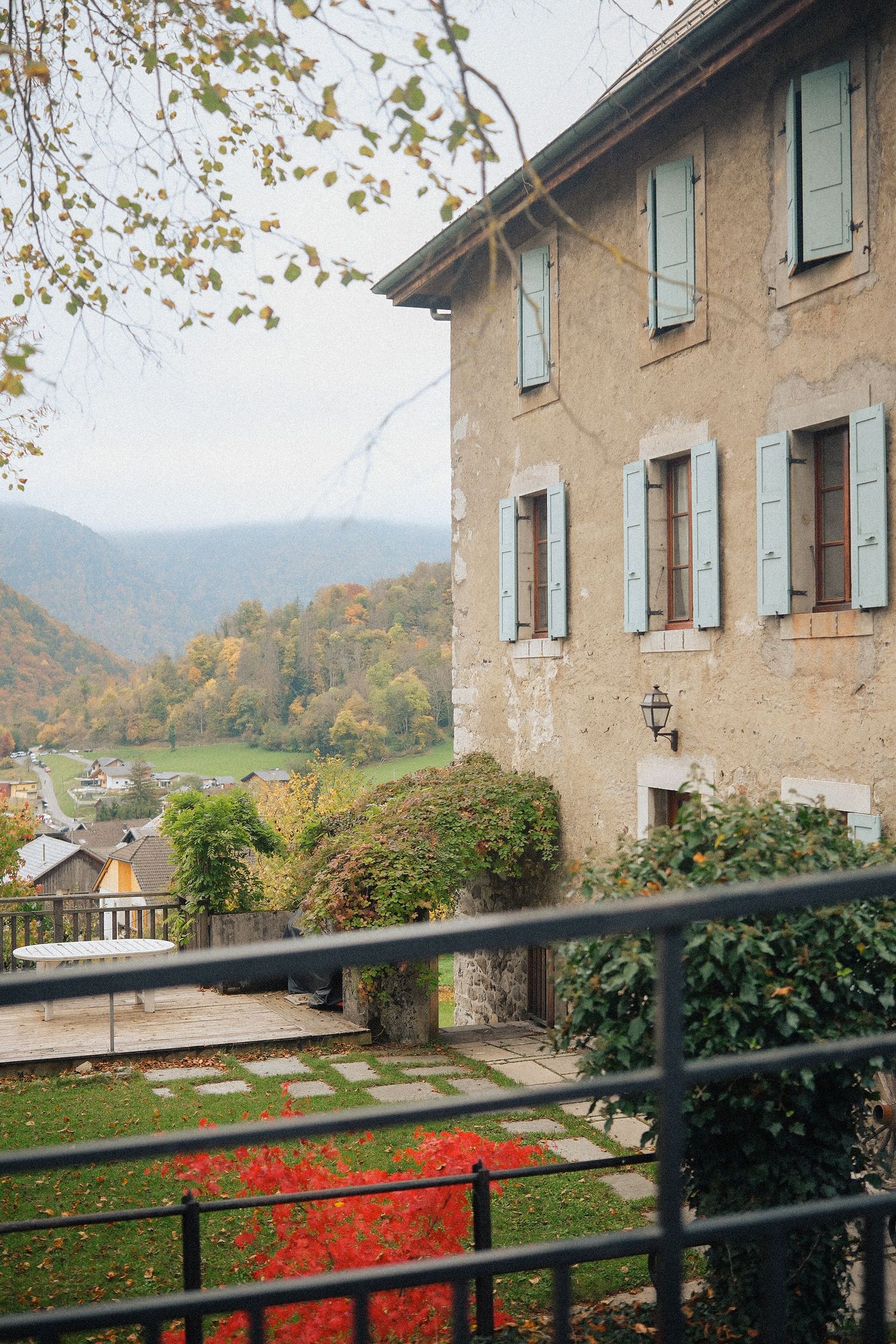 Autumn view of a rustic stone building with blue shutters overlooking a green valley and mountains.