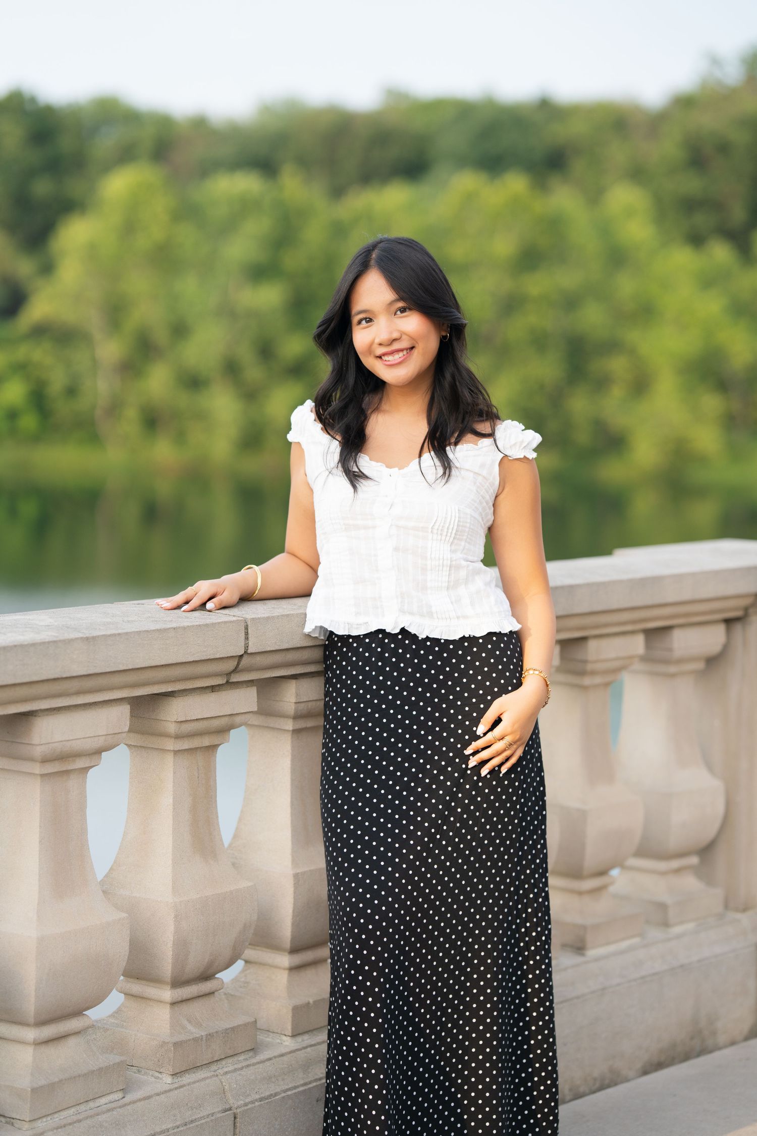 Young Asian woman in white top and polka dot maxi skirt smiling on stone balustrade with green lake backdrop.