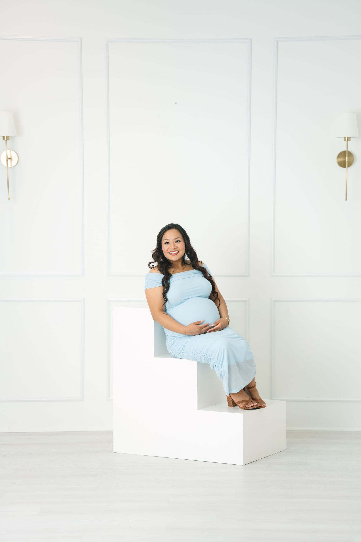 Pregnant woman in light blue dress sitting on white steps, cradling baby bump in bright white studio setting.
