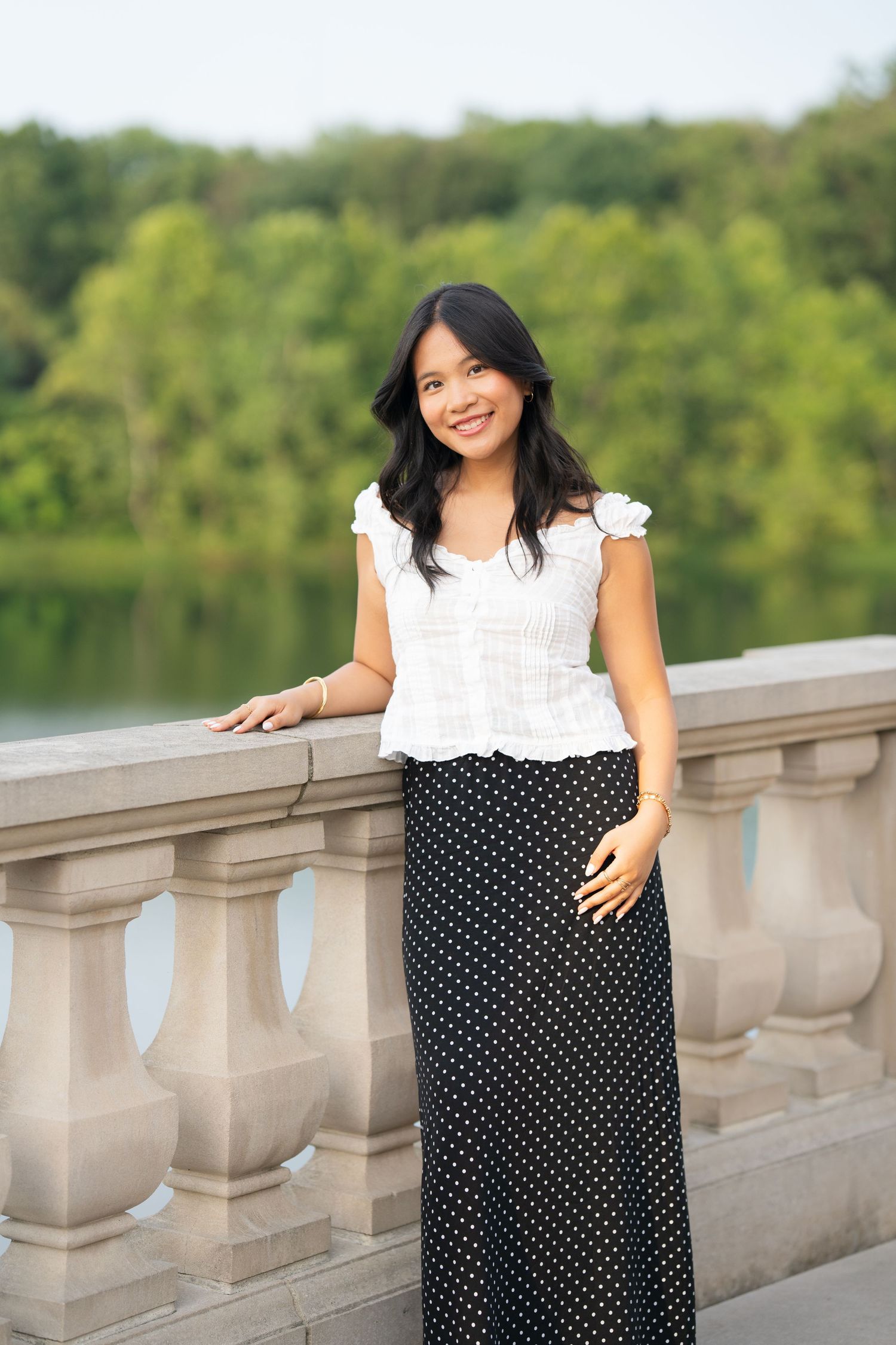 Young Asian woman in white top and polka dot maxi skirt posing on stone balustrade with green trees behind.