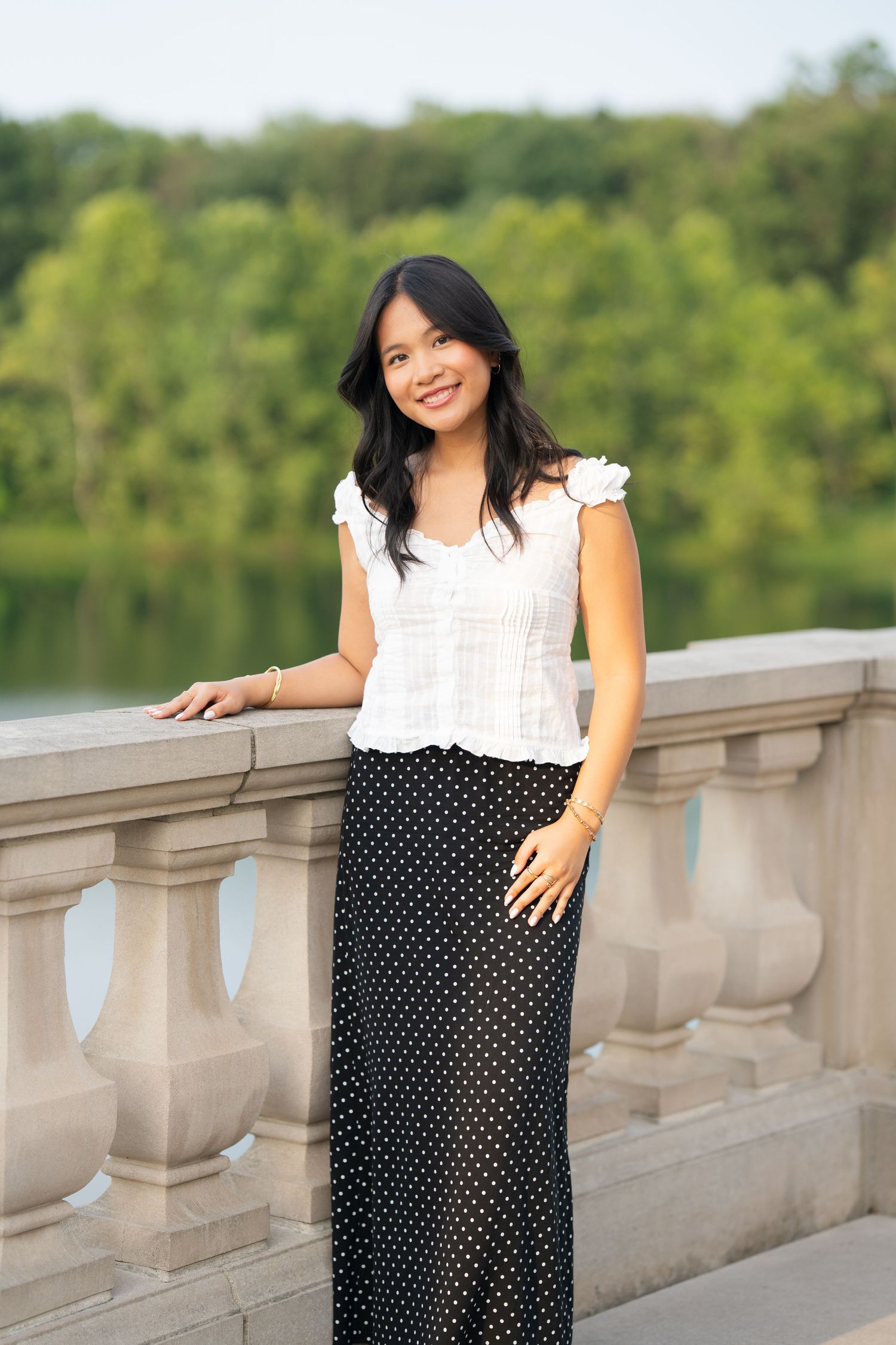 Young Asian woman in white top and polka dot maxi skirt smiling on stone balcony overlooking a serene lake.