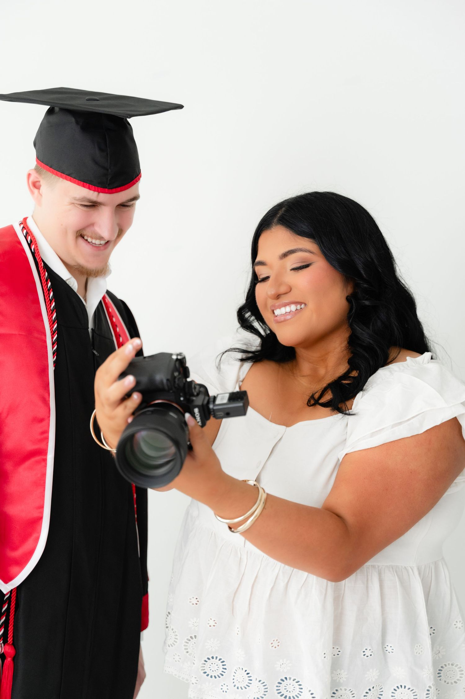 A graduate in cap and gown smiles while reviewing photos on a camera held by a woman in a white dress.