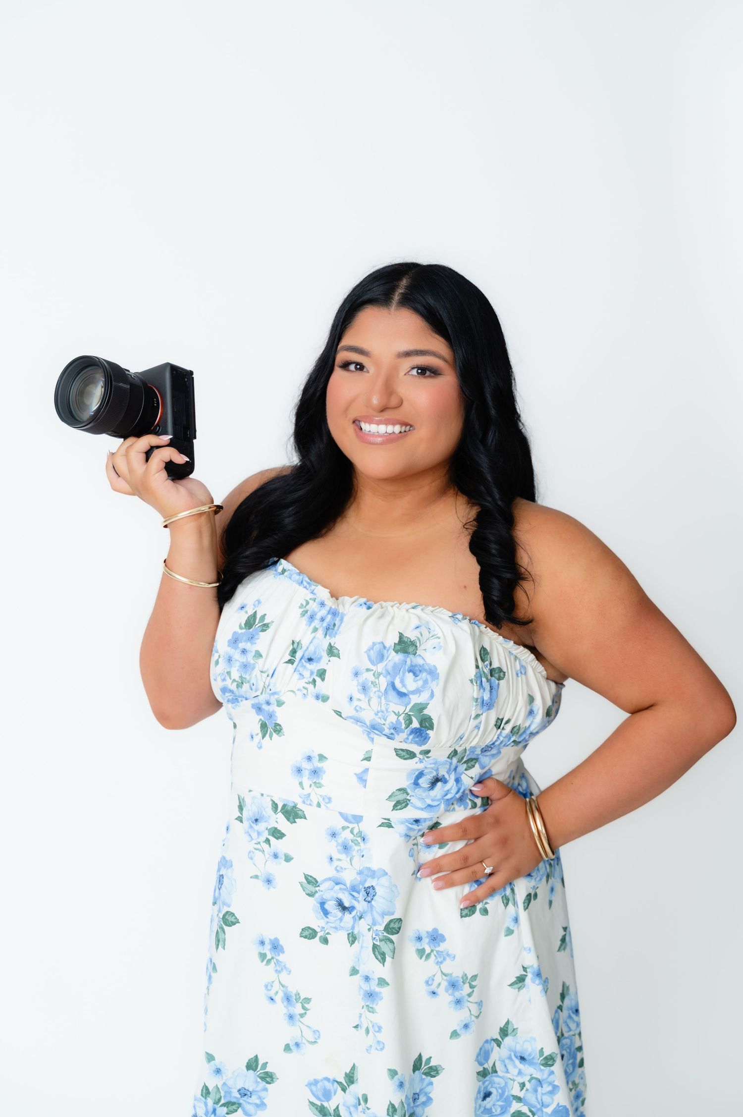 Smiling woman in floral blue dress holding a camera, posing against a white background.