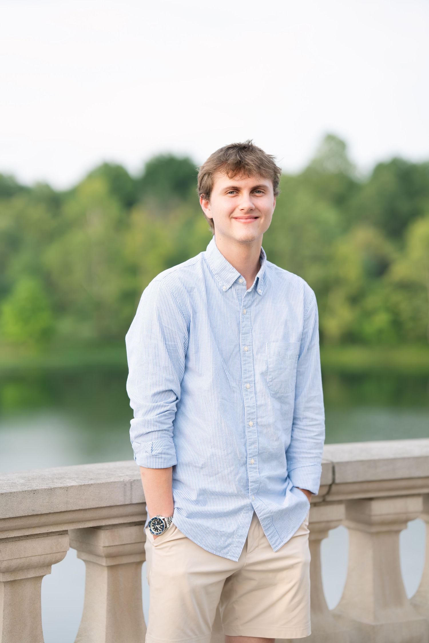 Young man in light blue shirt and khaki shorts smiling on a stone balcony with a lake and trees behind.