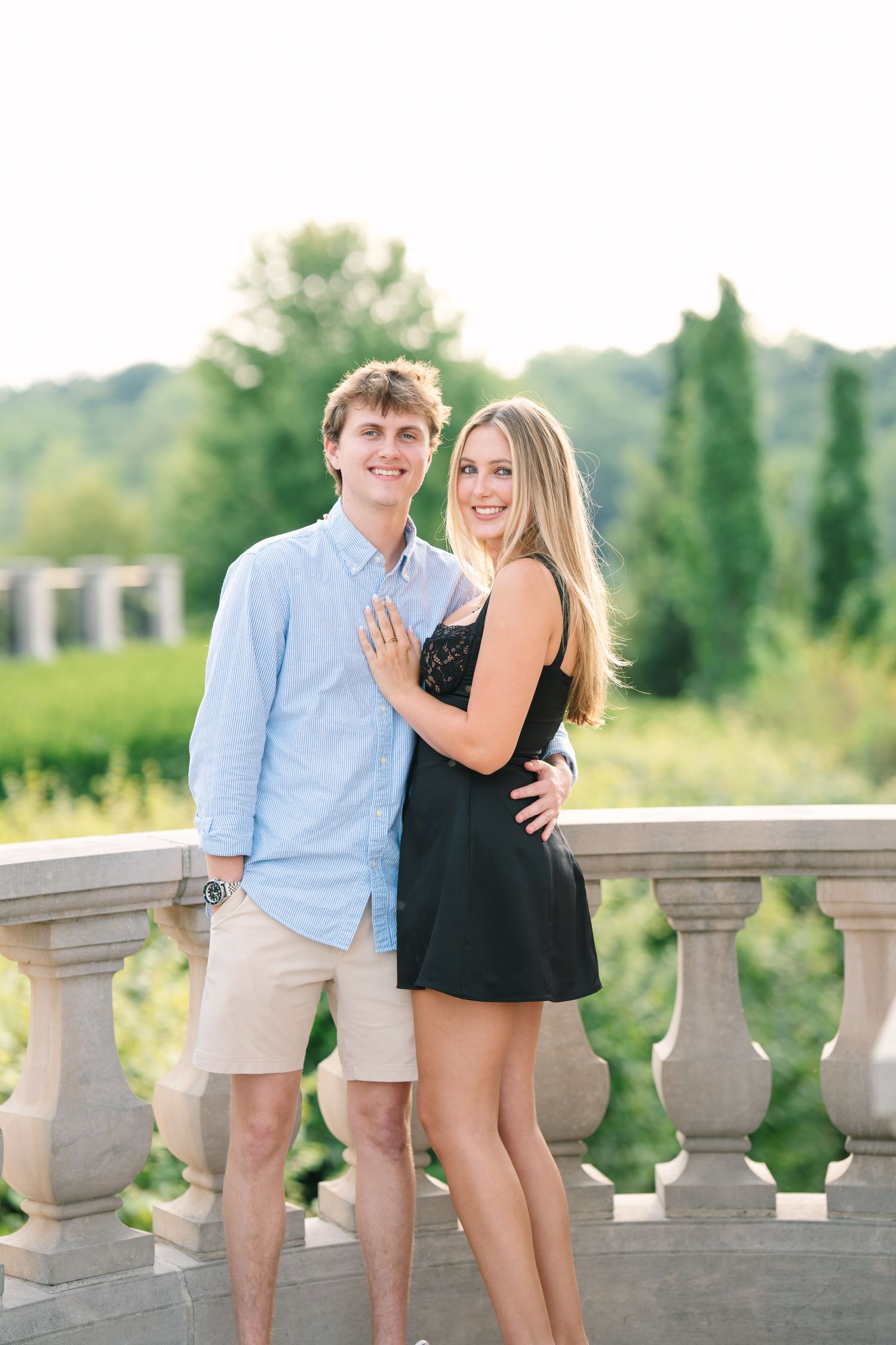 Smiling engaged couple posing on stone balcony at golden hour, woman showing off engagement ring.