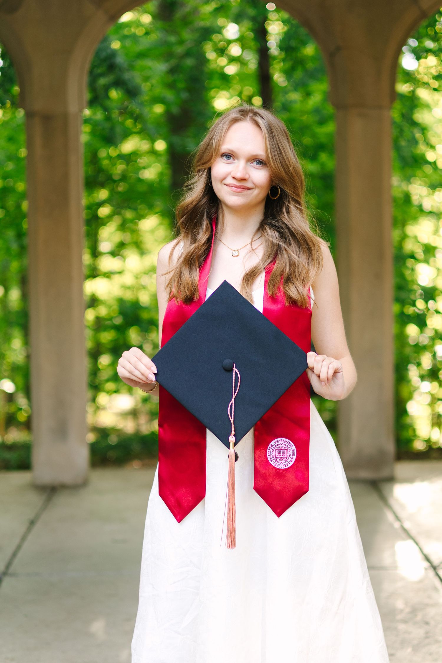 Young woman in white dress holding graduation cap with red tassel and stole, posing outdoors under stone arches.