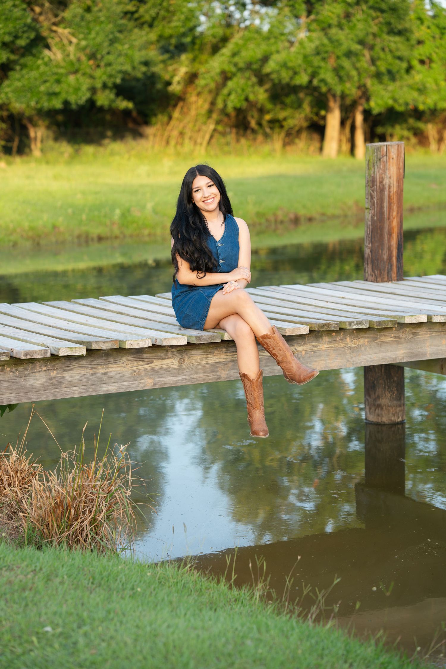 Young woman in blue dress and cowboy boots sitting on wooden dock over reflective pond.