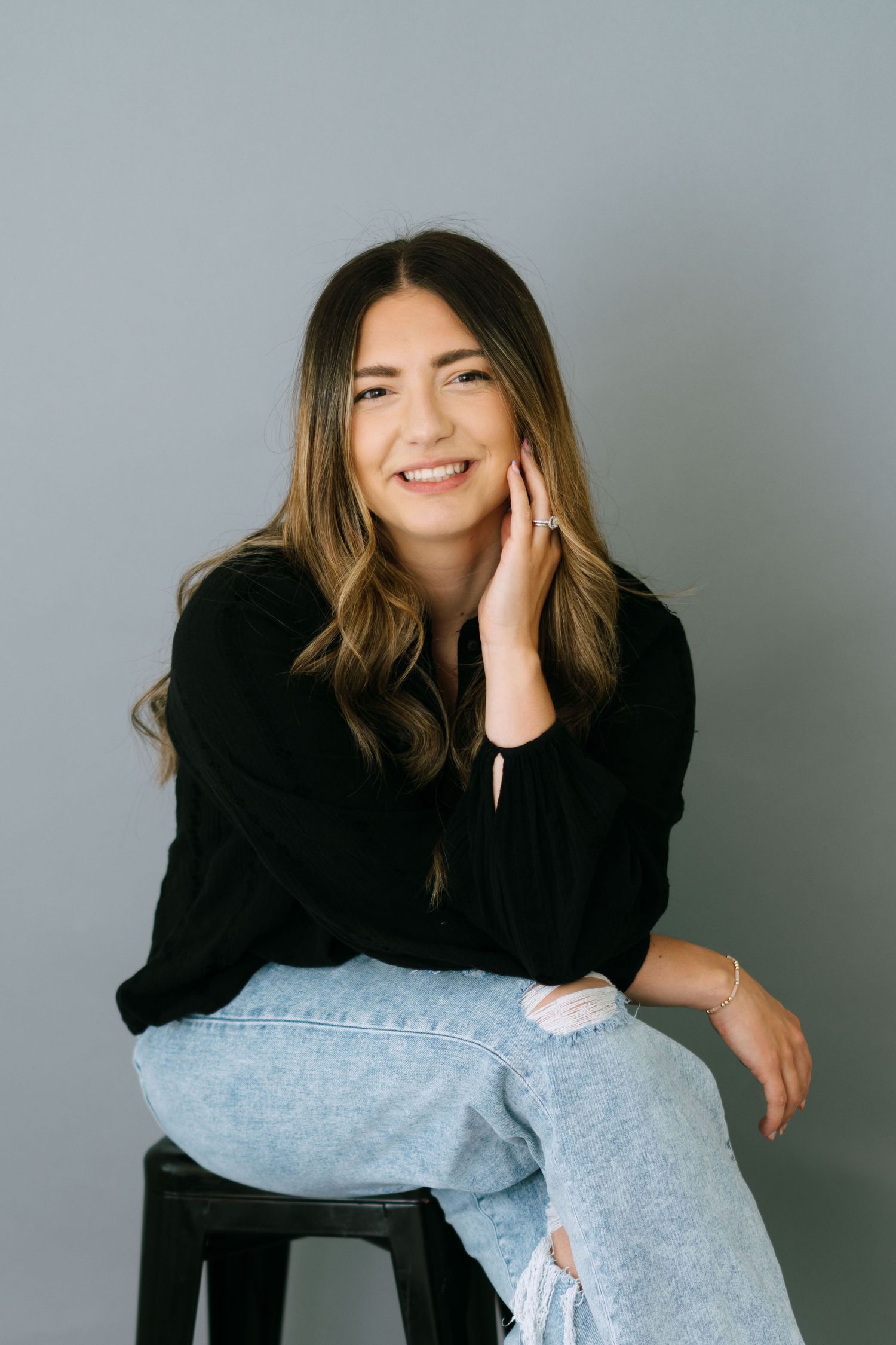Smiling young woman with highlighted hair sitting on a stool in a black top and ripped jeans.