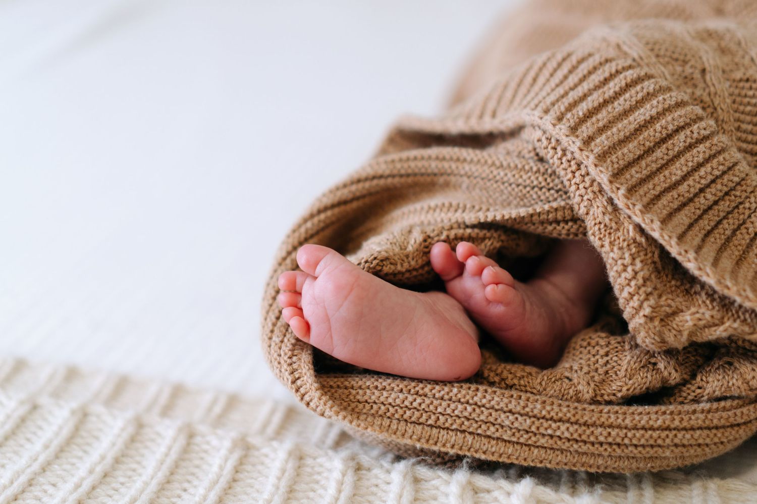 Tiny newborn baby feet peeking out from a cozy tan knit blanket on a soft white surface.