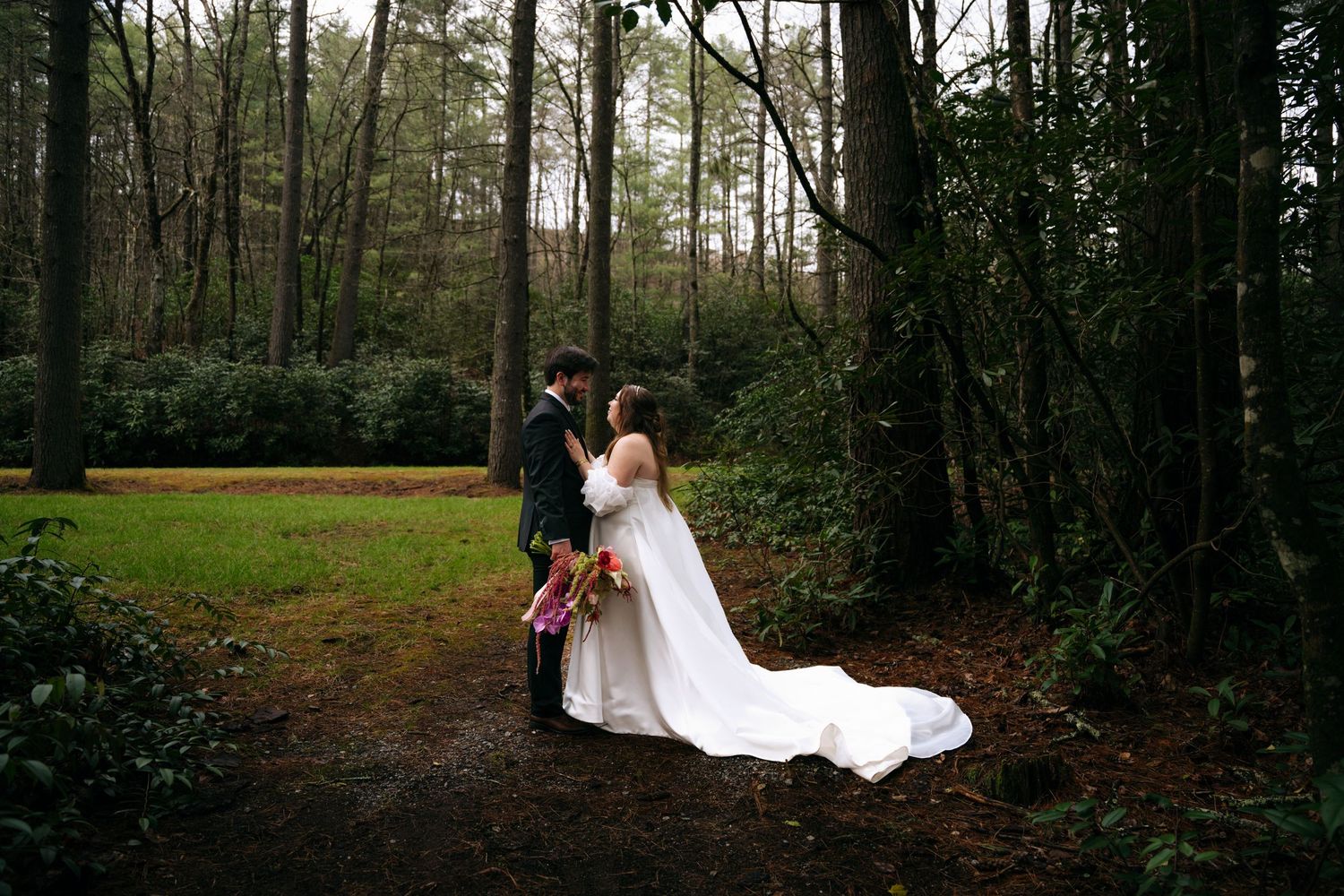 A bride and groom share a romantic moment on a forest path surrounded by tall trees.