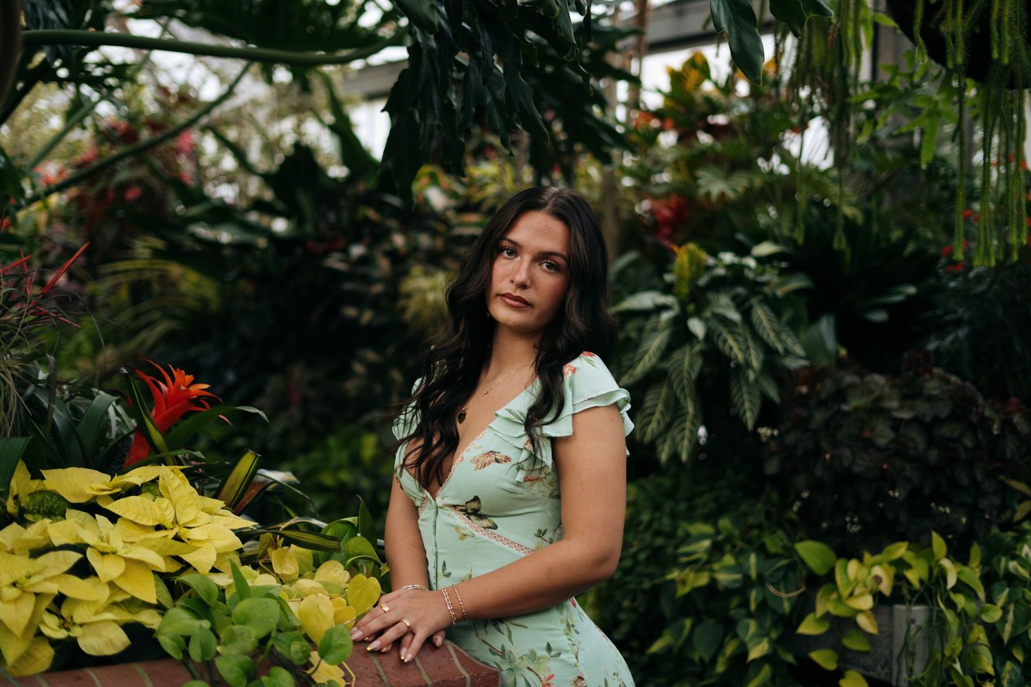 Young woman in mint floral dress posing gracefully among vibrant tropical plants in a greenhouse.