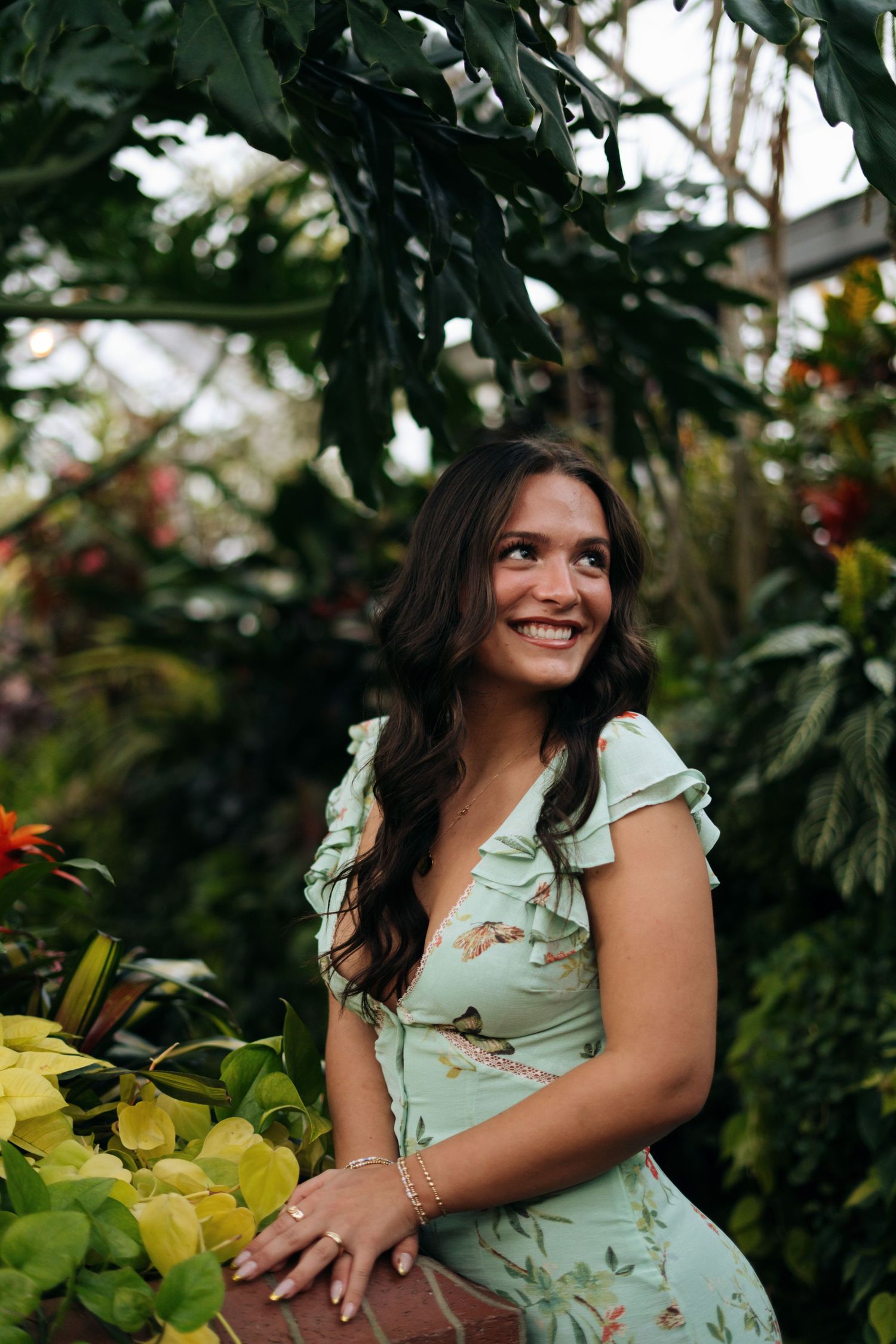 A smiling young woman in a mint floral dress sits among colorful tropical plants in a greenhouse.