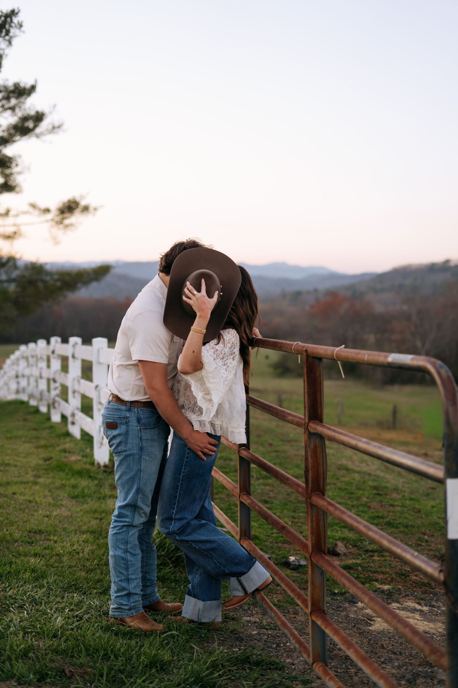 Couple sharing a kiss by a rustic farm gate at sunset with rolling green hills in the background.