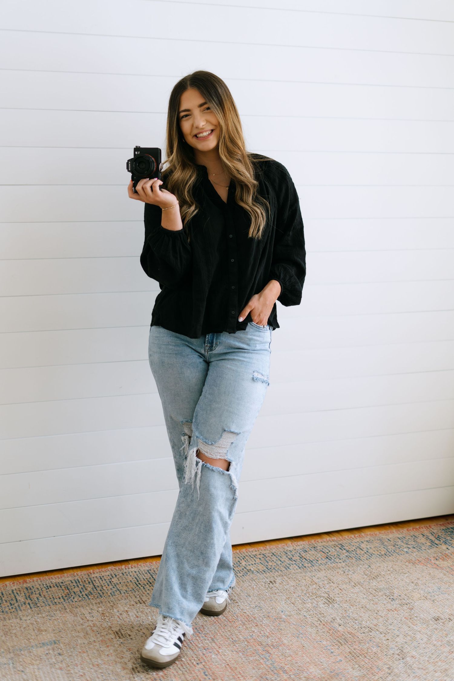 A smiling female photographer holds a camera while posing in a black shirt and ripped jeans.