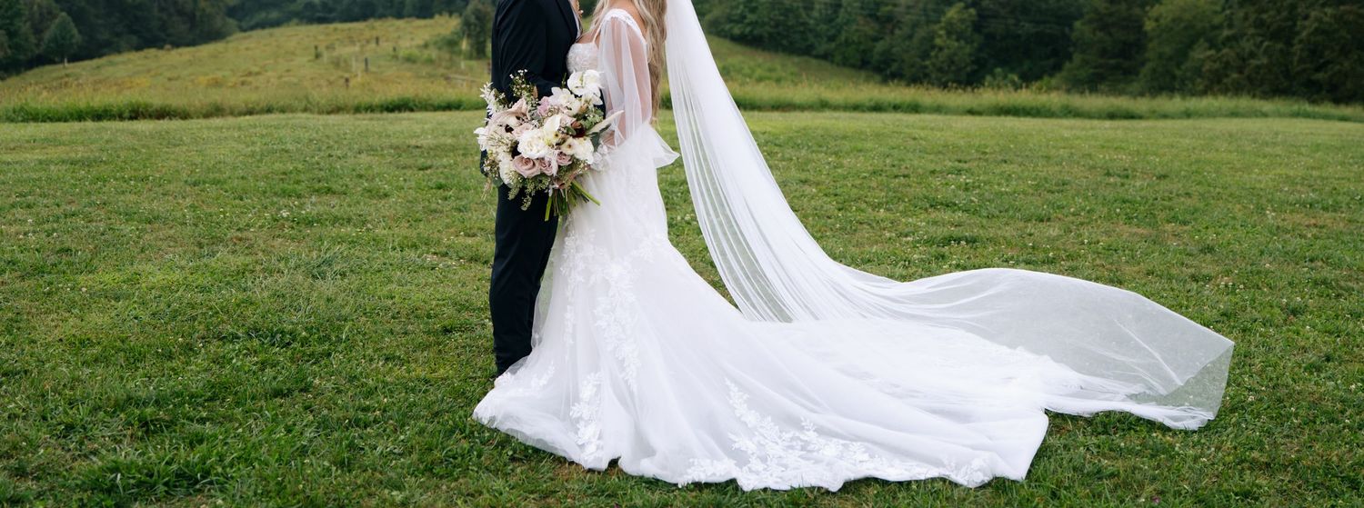 A bride in a flowing white gown with a long veil stands with a groom in a green meadow.