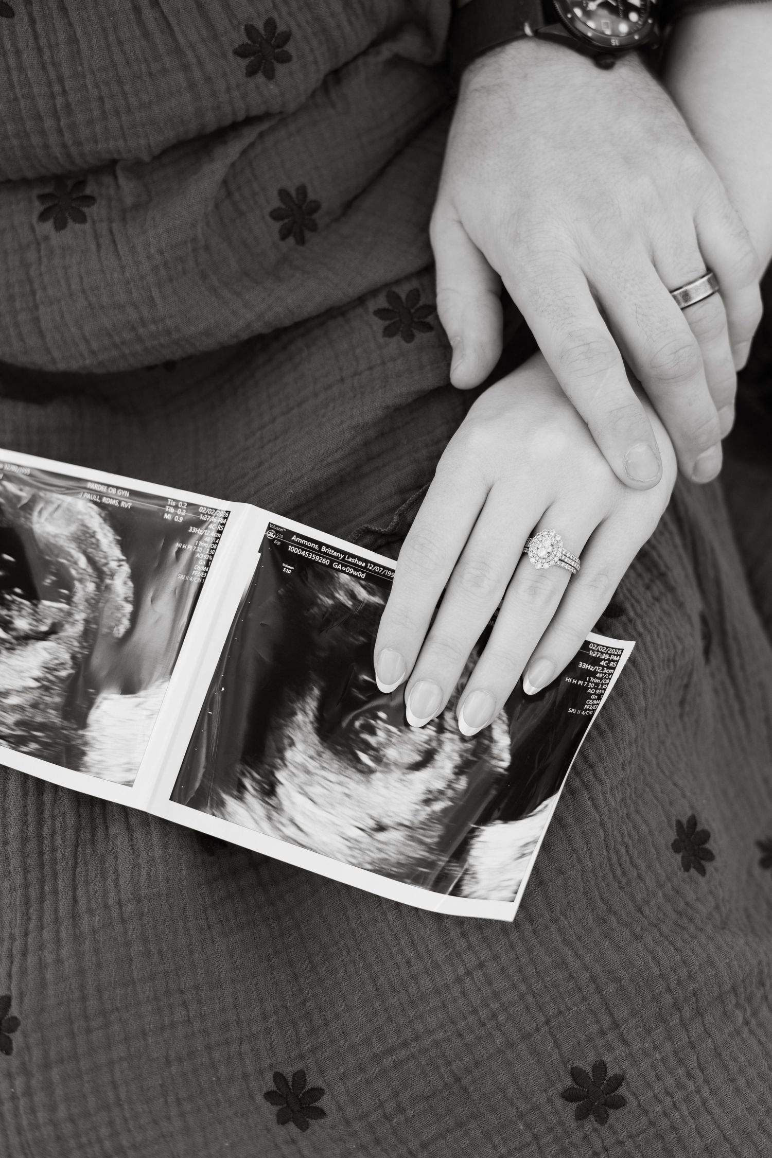 Black and white close-up of couple holding hands over ultrasound photos during pregnancy announcement.