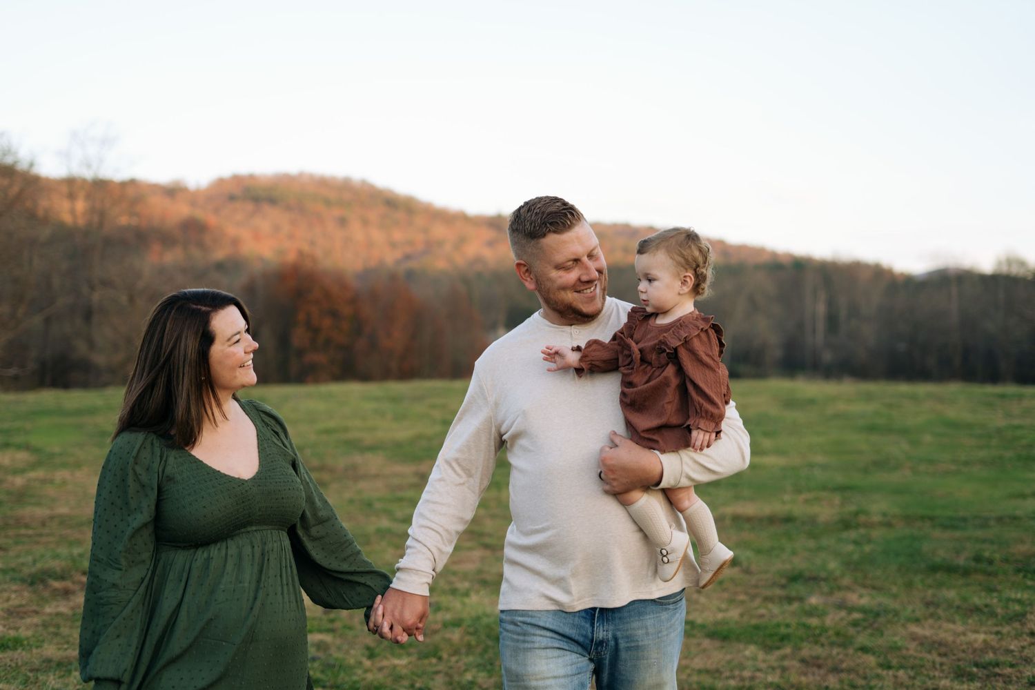 A happy family of three walks in a green field with mountains in the background during golden hour.