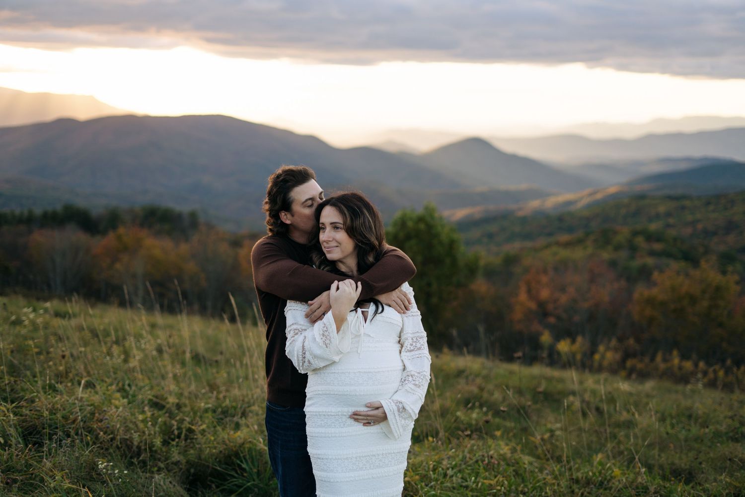 Couple embracing during maternity photoshoot at sunset with stunning mountain landscape backdrop.