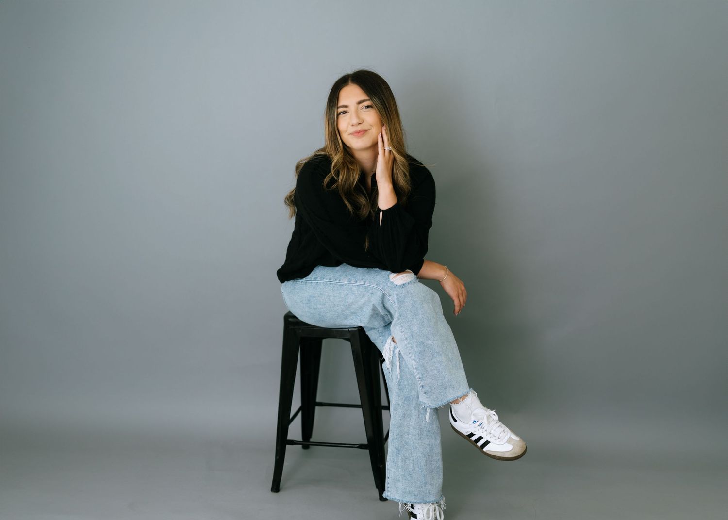 Young woman sitting on a stool in jeans and black top against gray background.