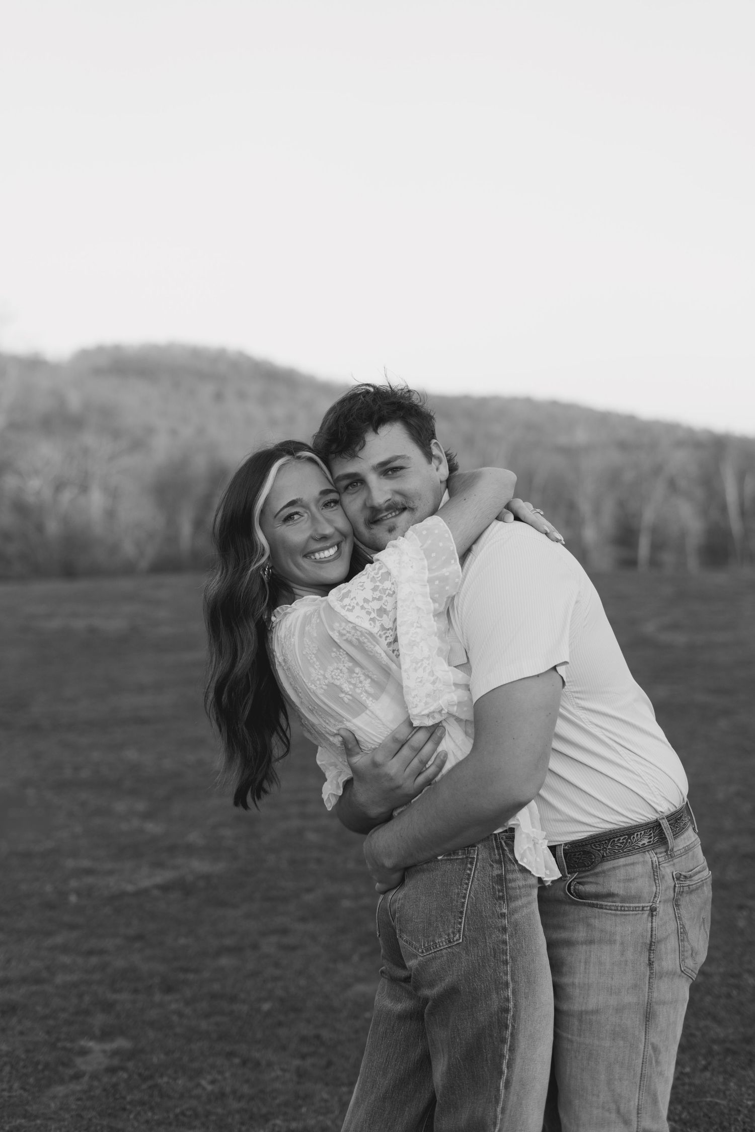 A smiling couple embraces in a black and white photo with rolling hills in the background.