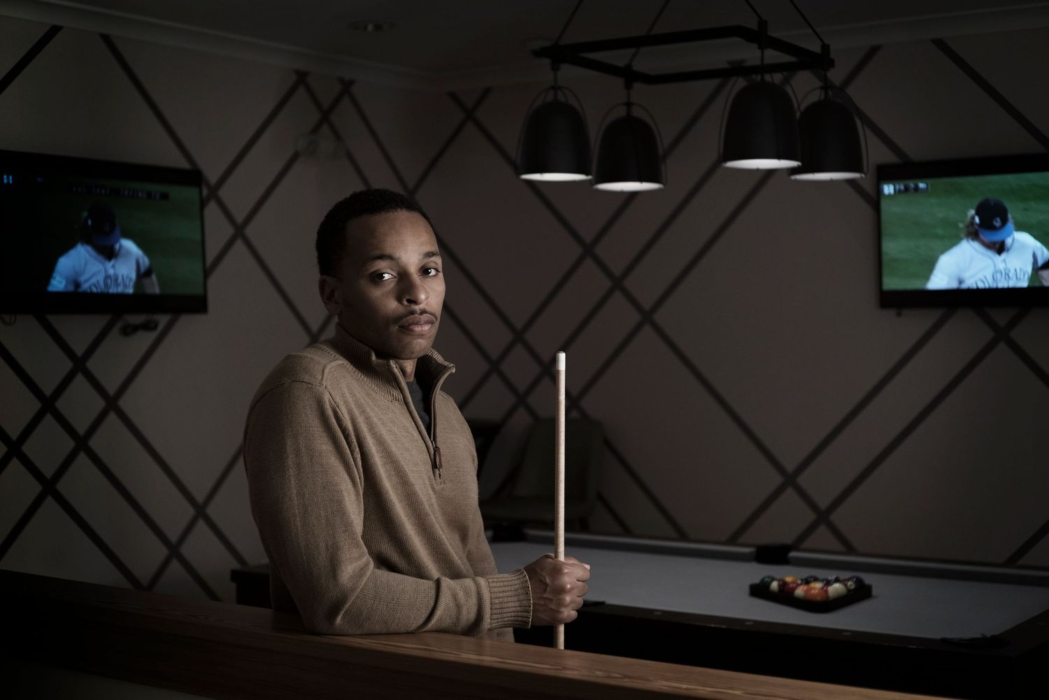 Man holding a pool cue beside a billiard table in a game room with TVs and pendant lighting.