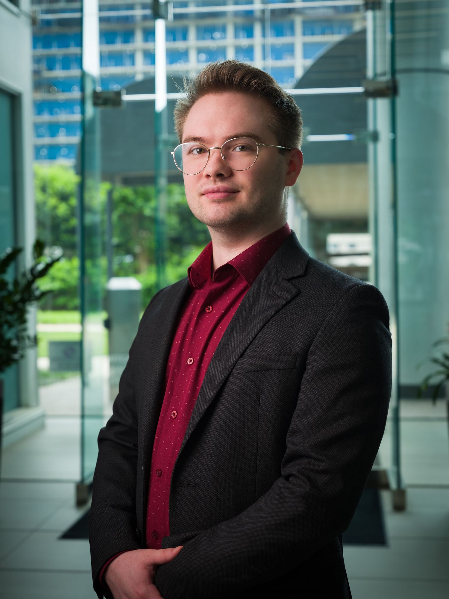 Young professional man in glasses and red shirt with black blazer posing in modern glass office building.