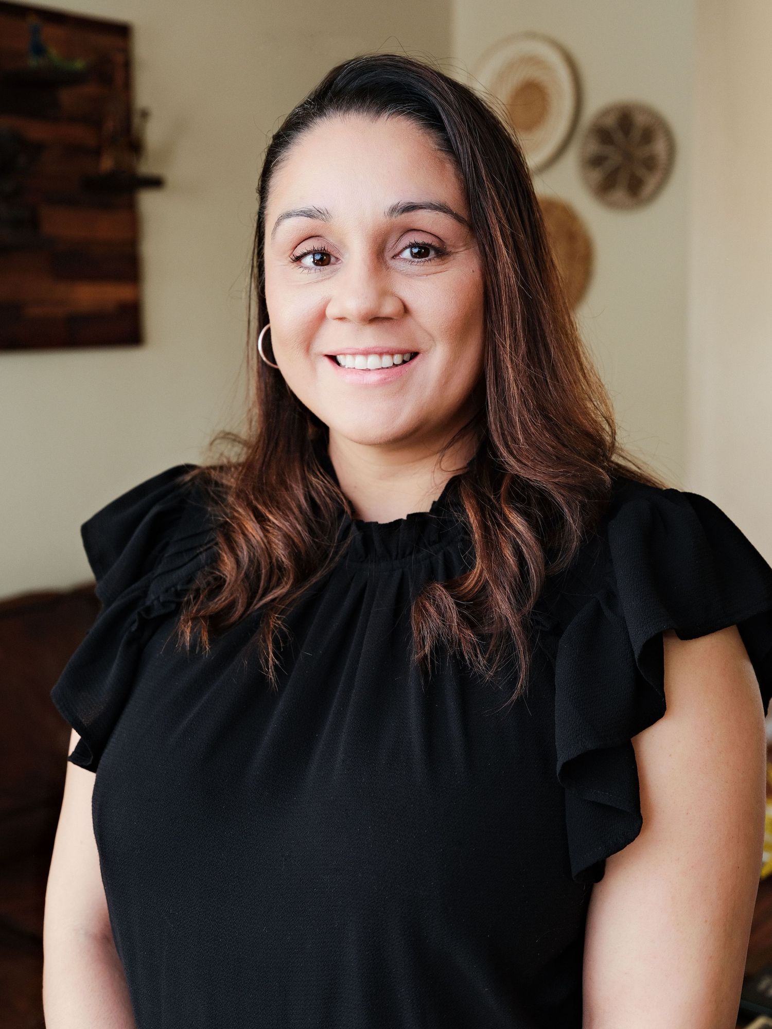 A smiling woman with long brown hair wearing a black ruffled top poses indoors for a professional headshot photo.