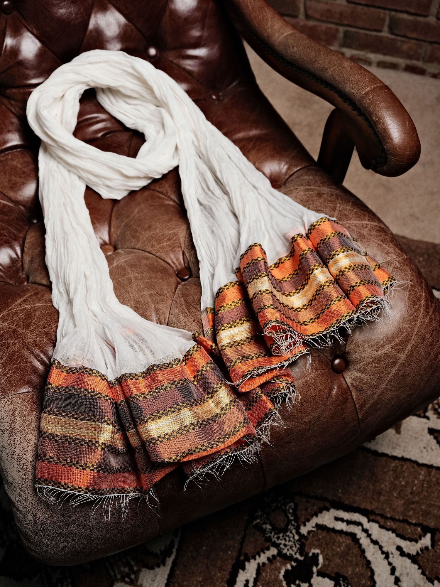 White and orange striped fringe scarf draped over a brown leather armchair.