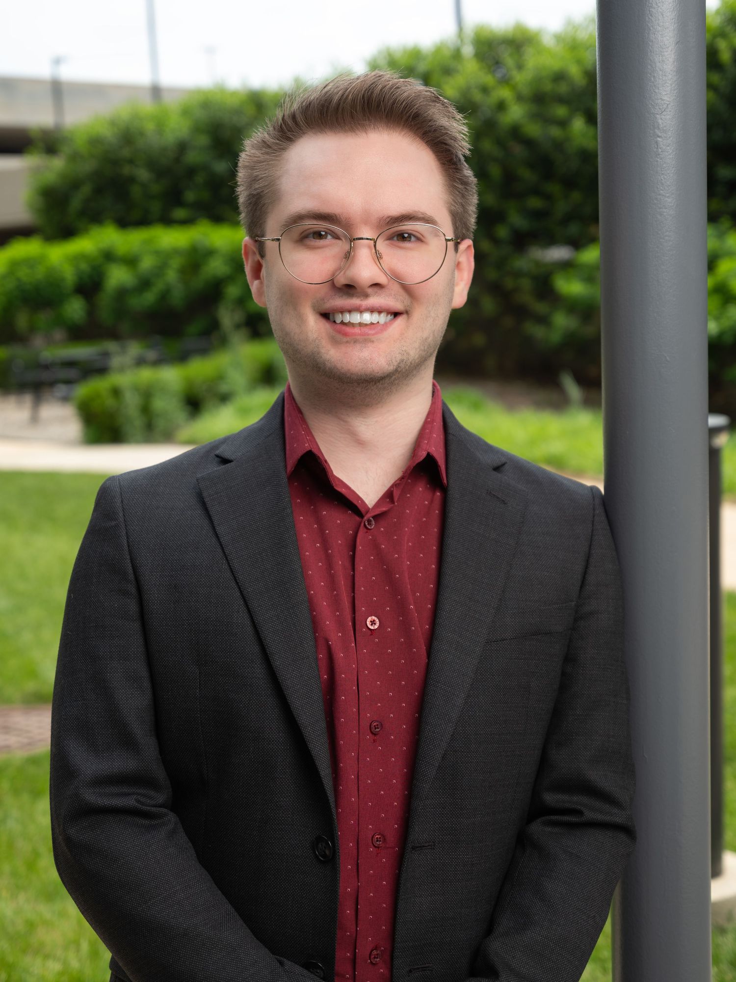 Young man in glasses and dark blazer smiling outdoors near greenery in a professional headshot photo.