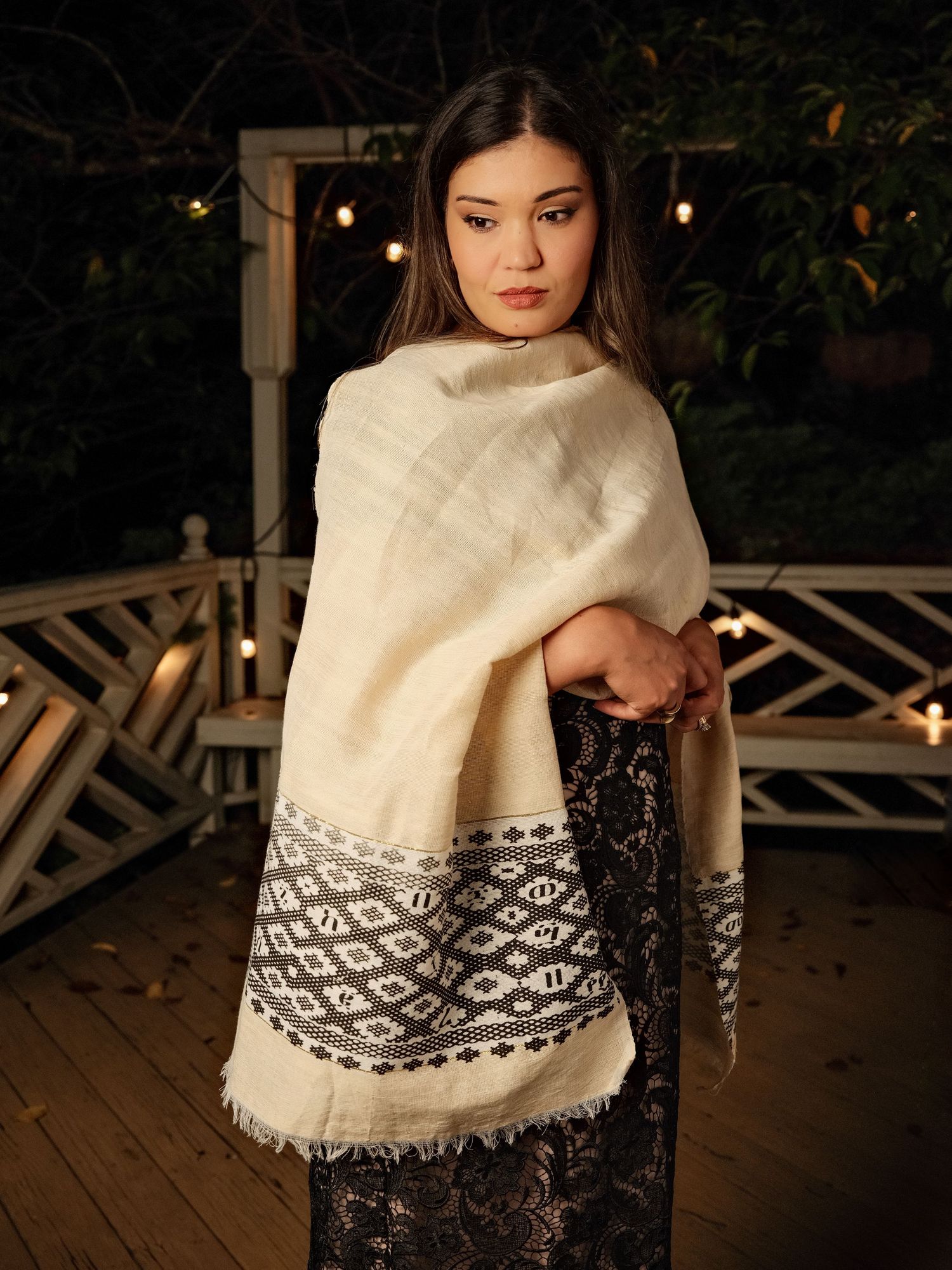 Woman wearing a cream shawl with black geometric border pattern, draped elegantly over shoulders on a wooden deck at night.
